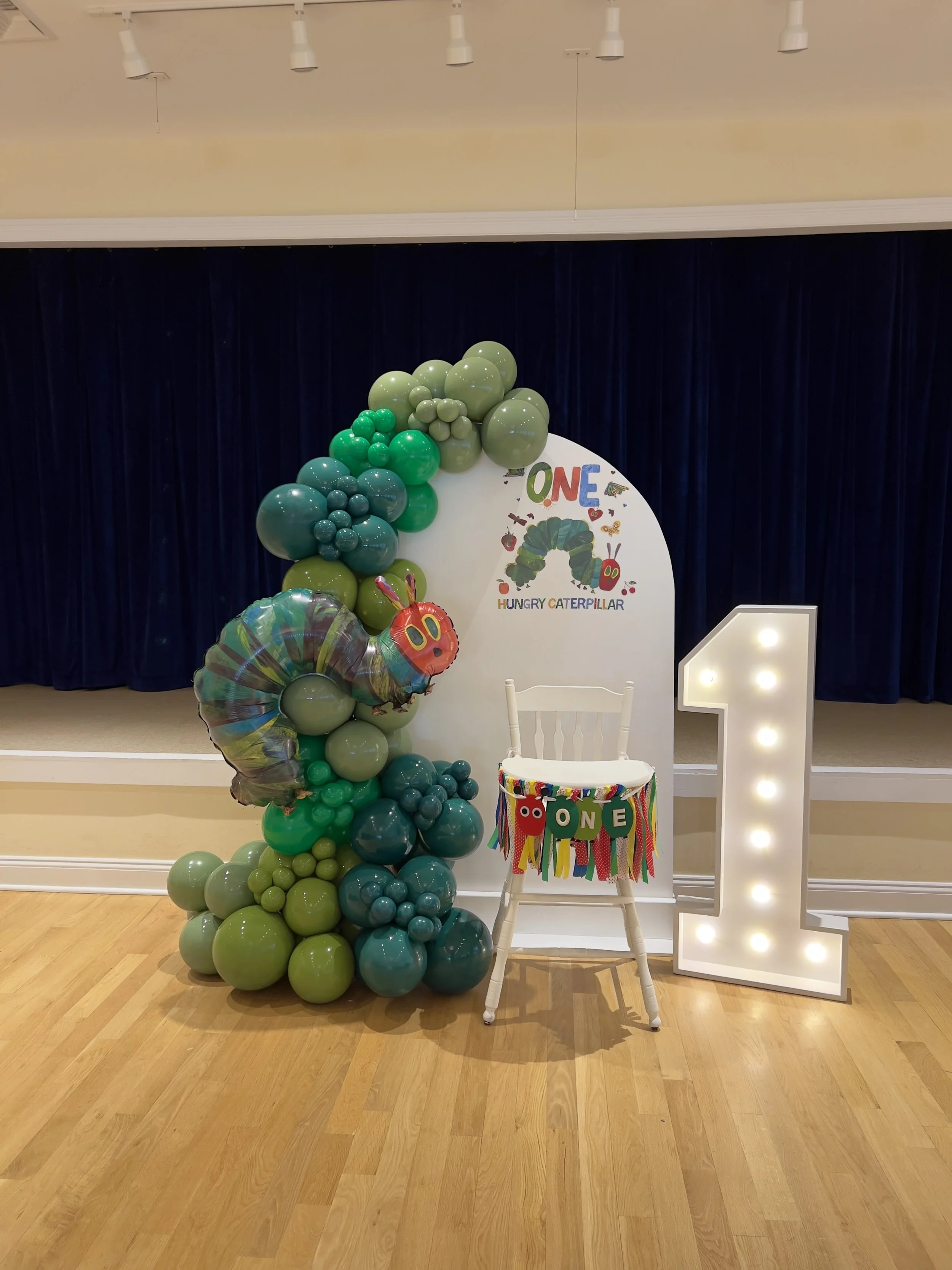 First birthday party decoration with a large illuminated number one, balloons, and a sign featuring a cartoon caterpillar, set against a dark blue curtain background.
