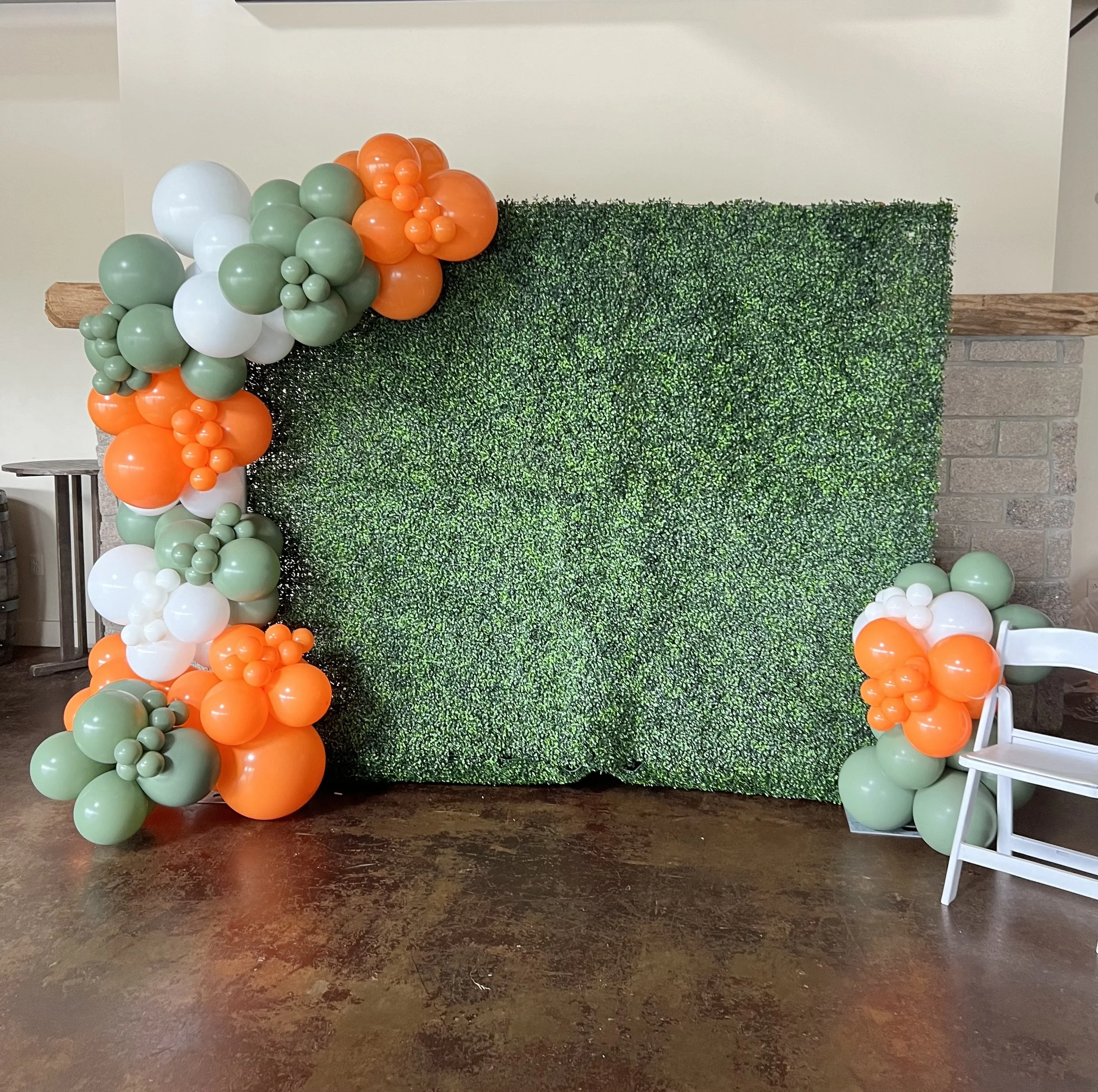 Decorative wall with green foliage backdrop, accented with balloons in orange, white, and green, arranged on the left and right sides.