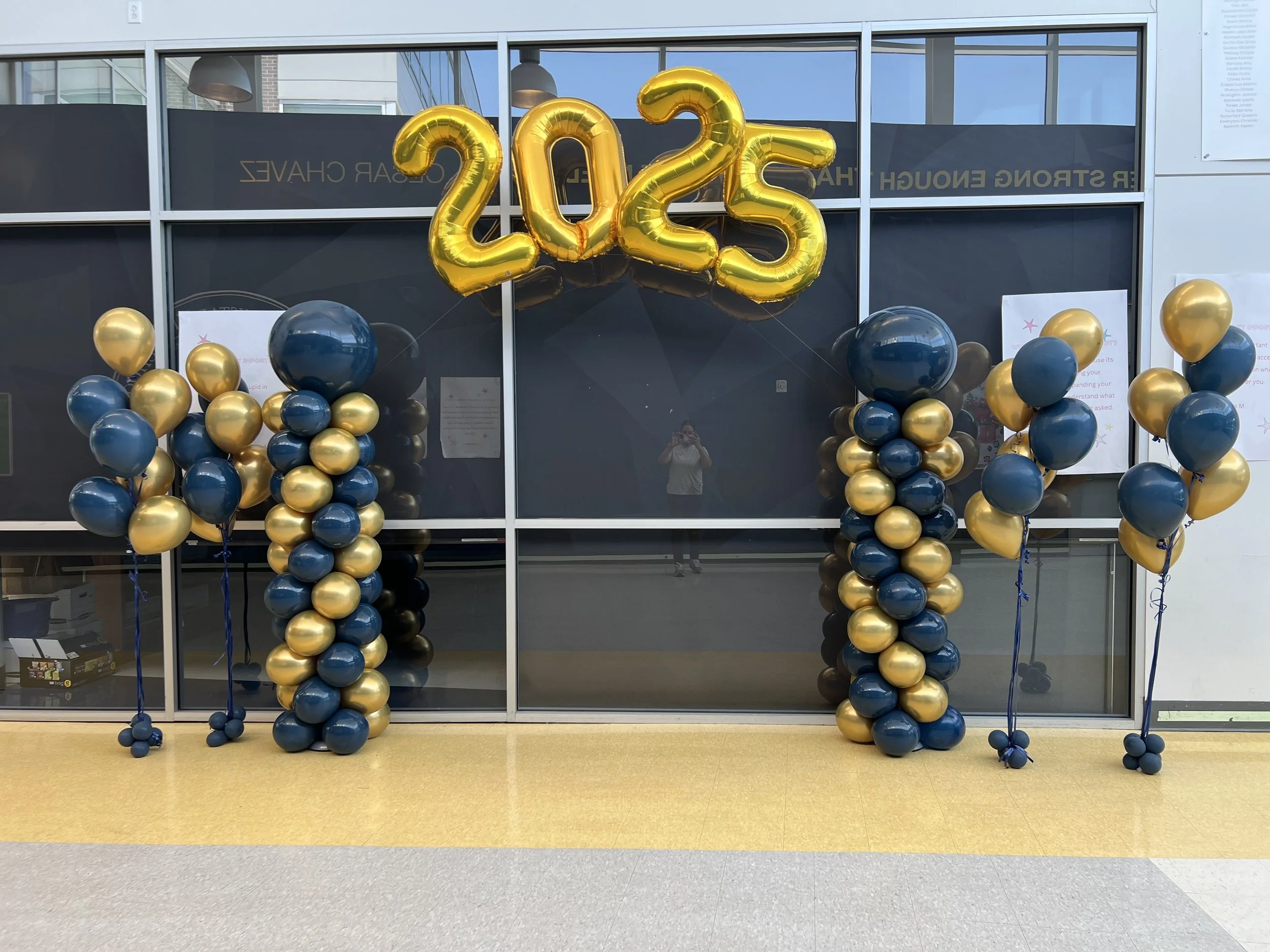 Decorative balloons with blue and gold colors arranged in columns, black and gold balloons form the number and letter at the top in gold, on the exterior of a modern glass-front building. The balloons spell out '2025' in large gold foil numbers and l
