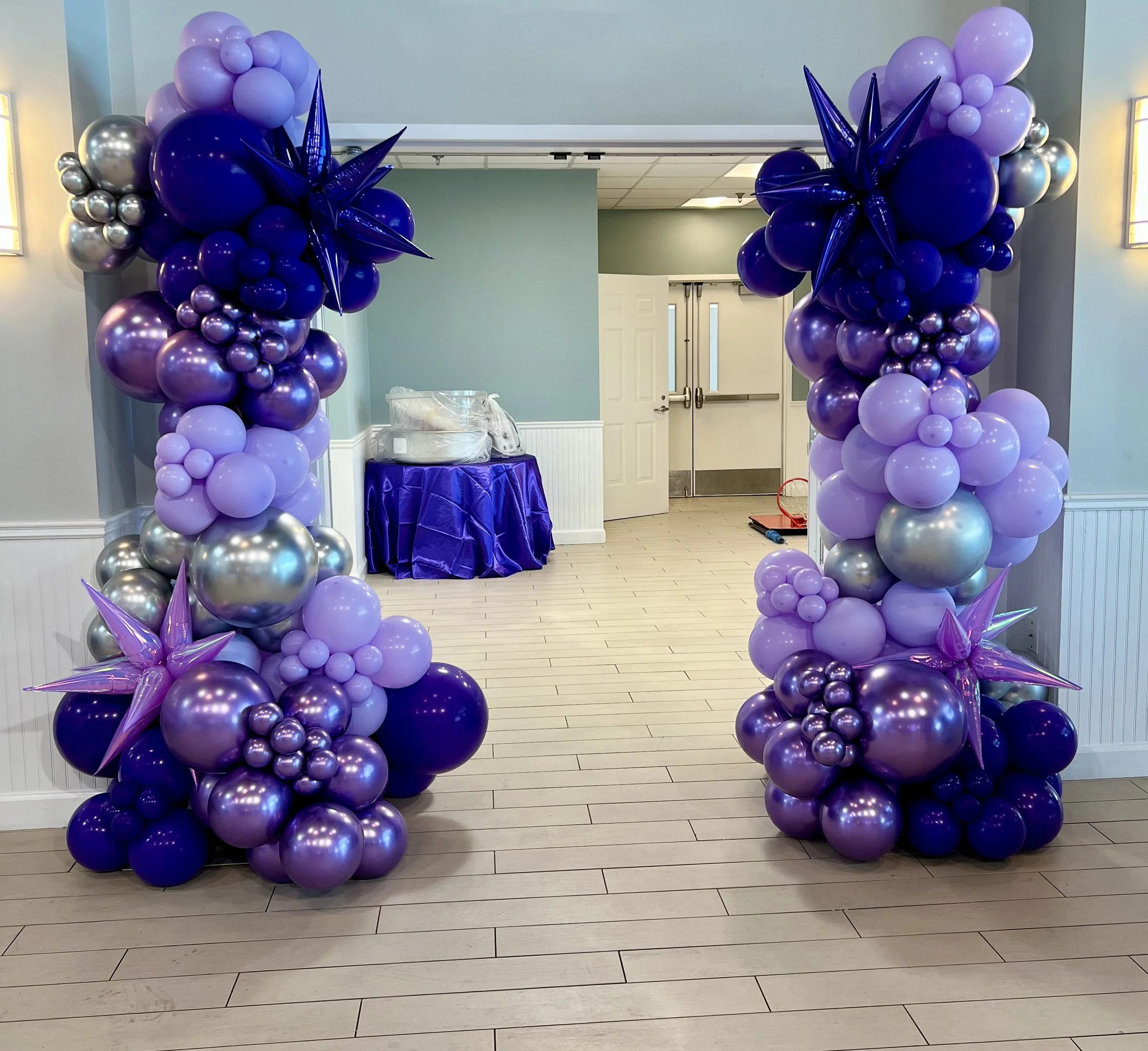 Decorative balloon arch in shades of purple, lavender, and silver with star-shaped balloons at the entrance of an event space, featuring balloons clustered at the base and balloons shaped as stars at the top.