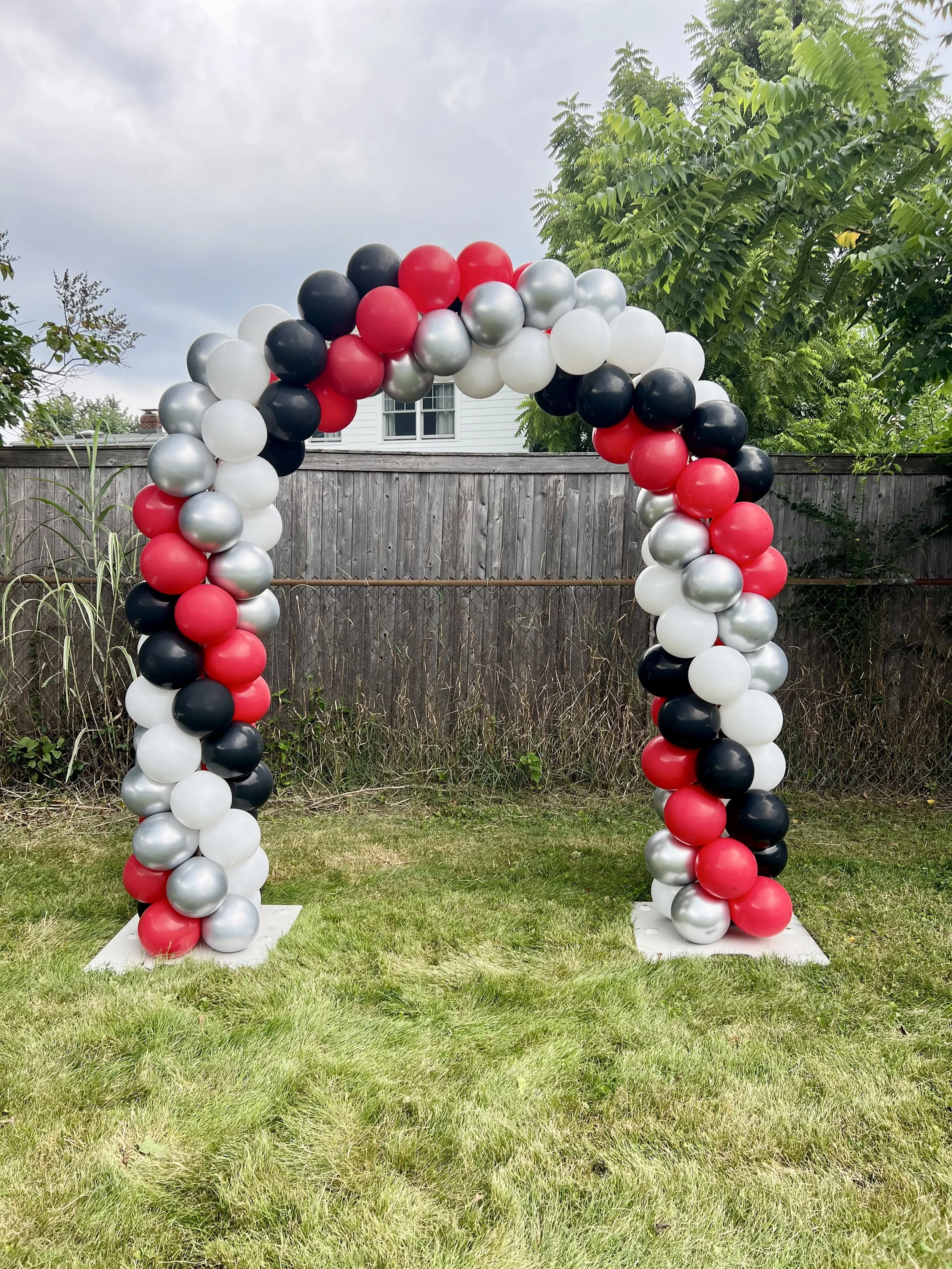 Balloon arch in red, black, white, and silver colors on a grassy backyard with a wooden fence and trees.