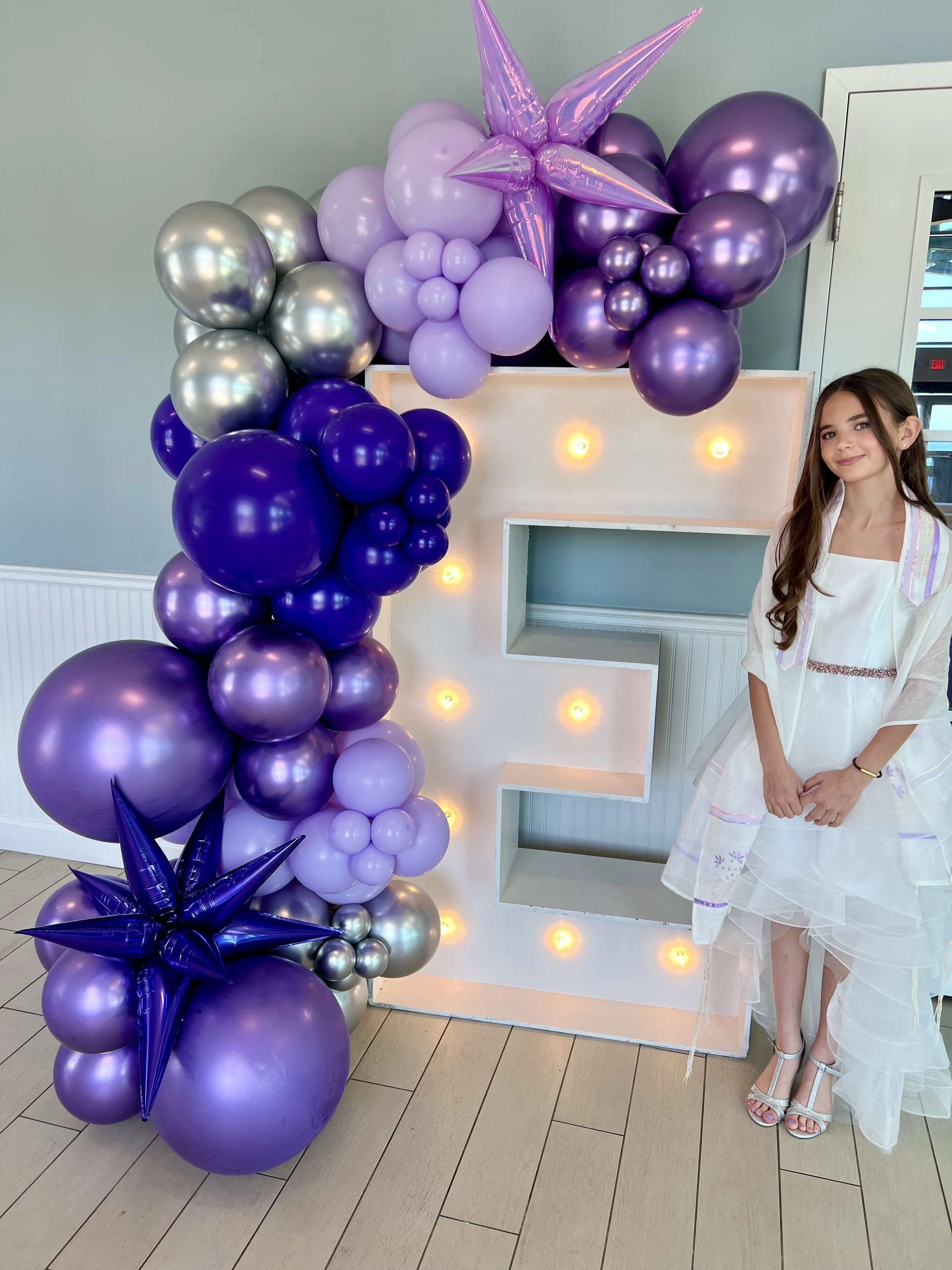 A young girl in a white dress standing next to a large decorative lighted letter E with purple, lavender, and silver balloons arranged around it, some shaped like stars, at a celebration or party.