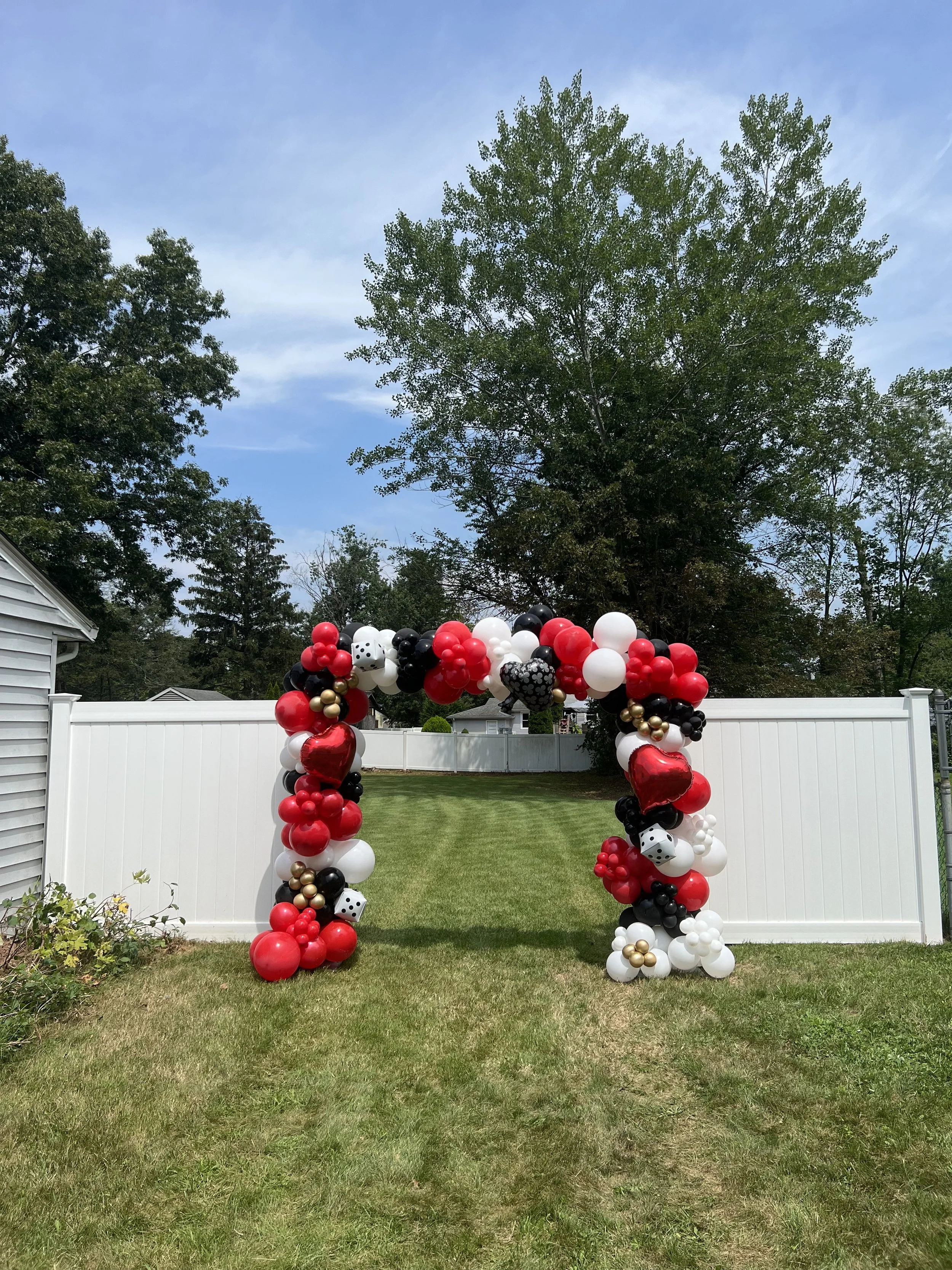 Decorative balloon arch with red, black, white, and gold balloons in a backyard with green grass and white fence.