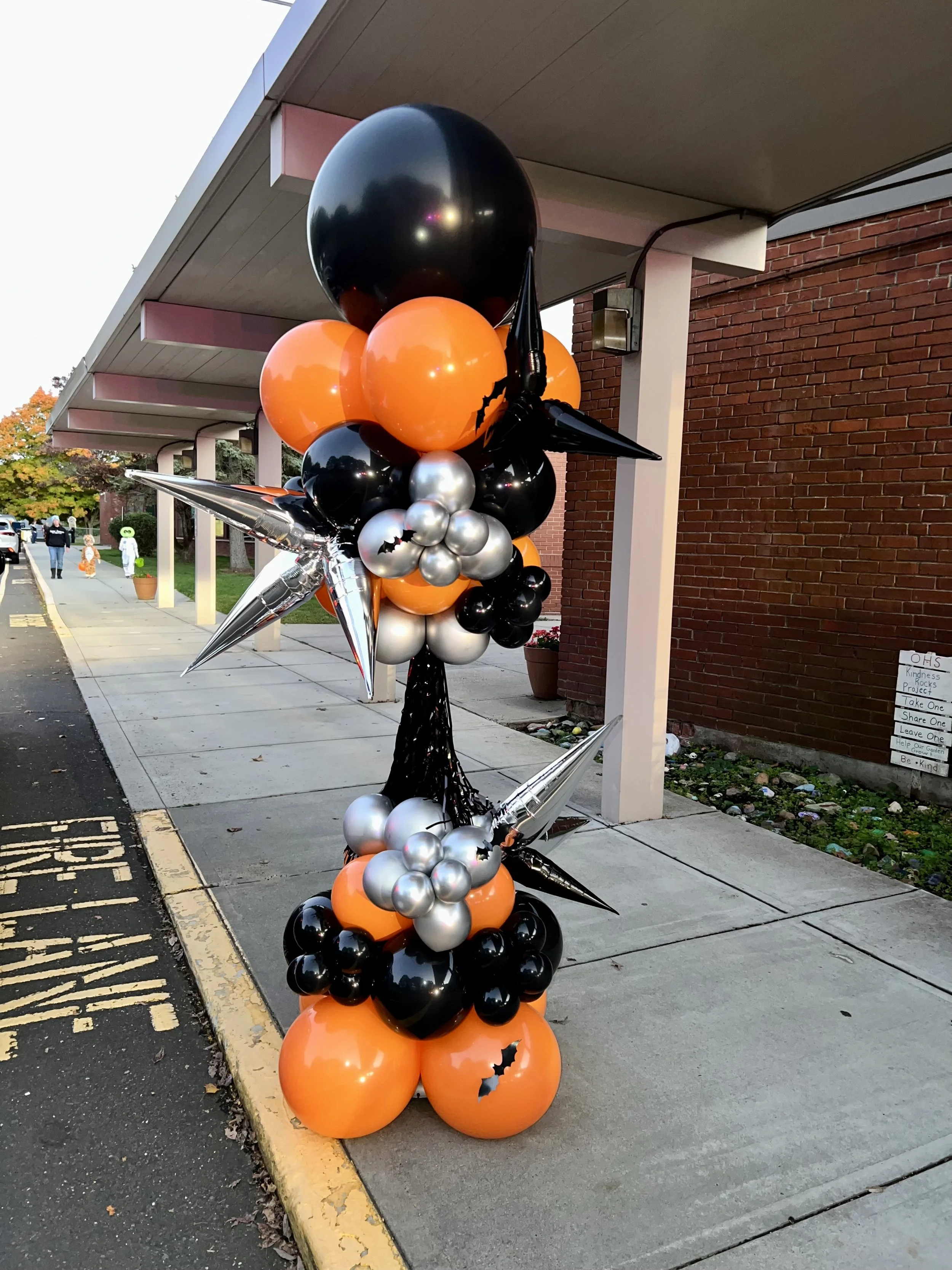 Halloween-themed balloon decoration with black, orange, silver, and metallic balloons, shaped with stars and bats, placed outside a building on the sidewalk.