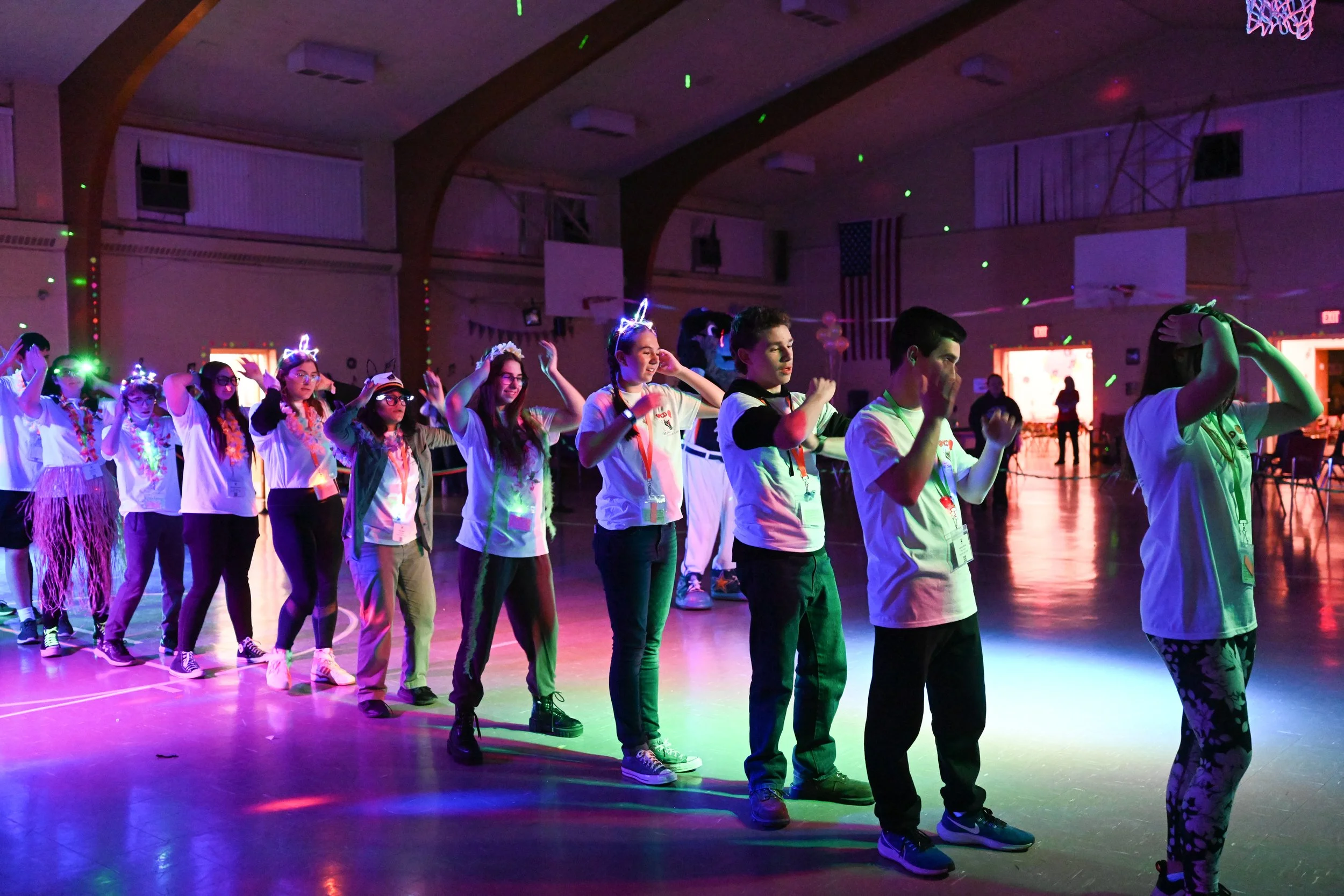 A group of children at a party or dance event inside a gymnasium, wearing glow-in-the-dark headbands and leis, standing in a line and dancing under colorful lighting.