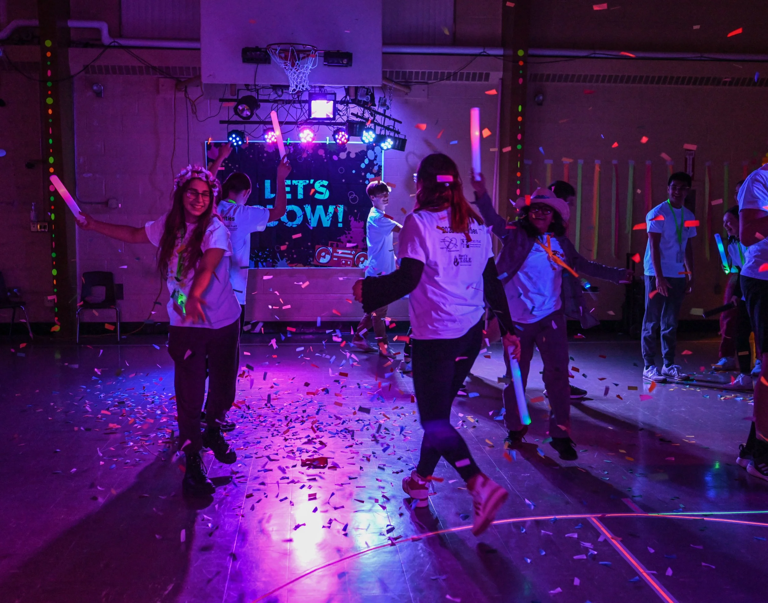 People dancing at a party or celebration with colorful lights, confetti, and a sign that says "Let's Glow" in the background.