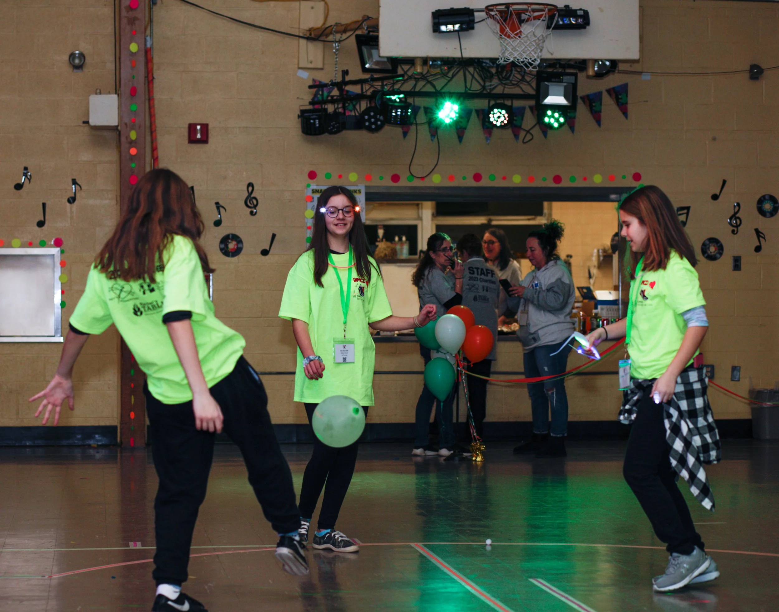 Three girls in neon green shirts playing a game with balloons and glow sticks in a decorated gymnasium with a DJ setup and audience in the background.