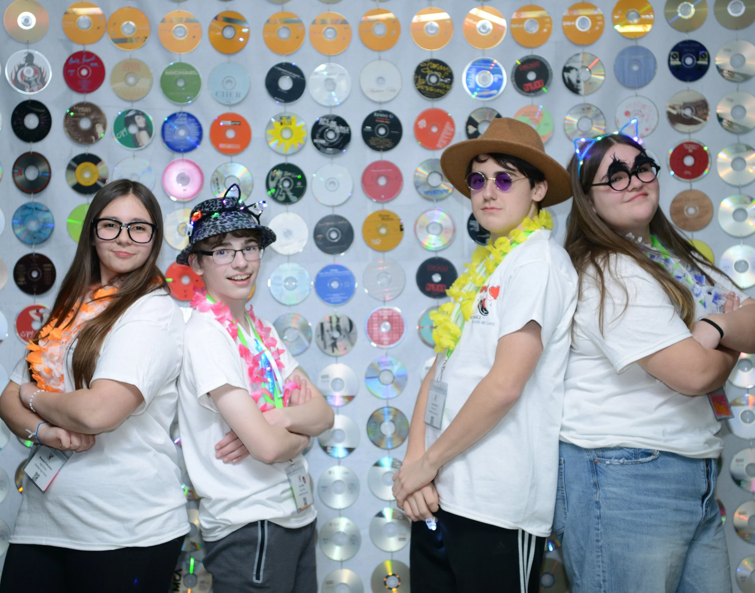 Four young women standing in front of a wall decorated with numerous CDs, wearing casual clothes, party accessories, and leis, smiling and posing for the photo.