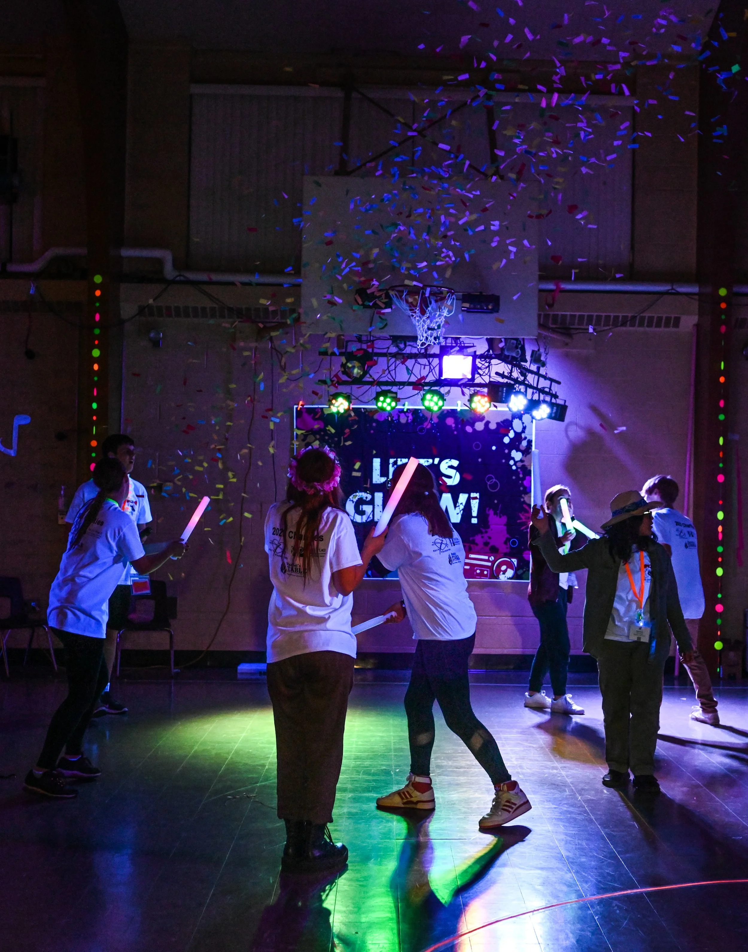 A group of people celebrating in a gymnasium with colorful lights, confetti, and a basketball hoop, with a sign that says "Let's Glow!"