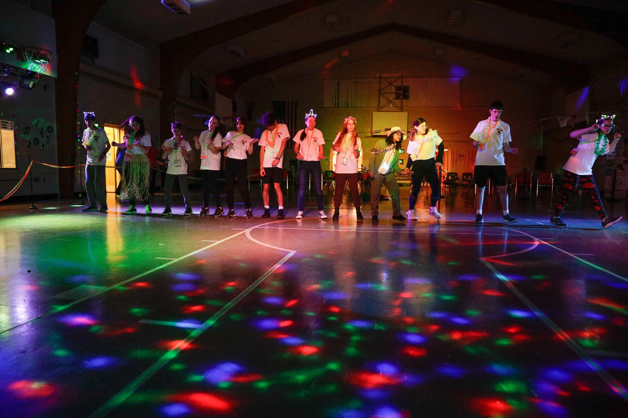 Children and teenagers standing in a line on a gymnasium floor with colorful disco lights, possibly during a dance or party.