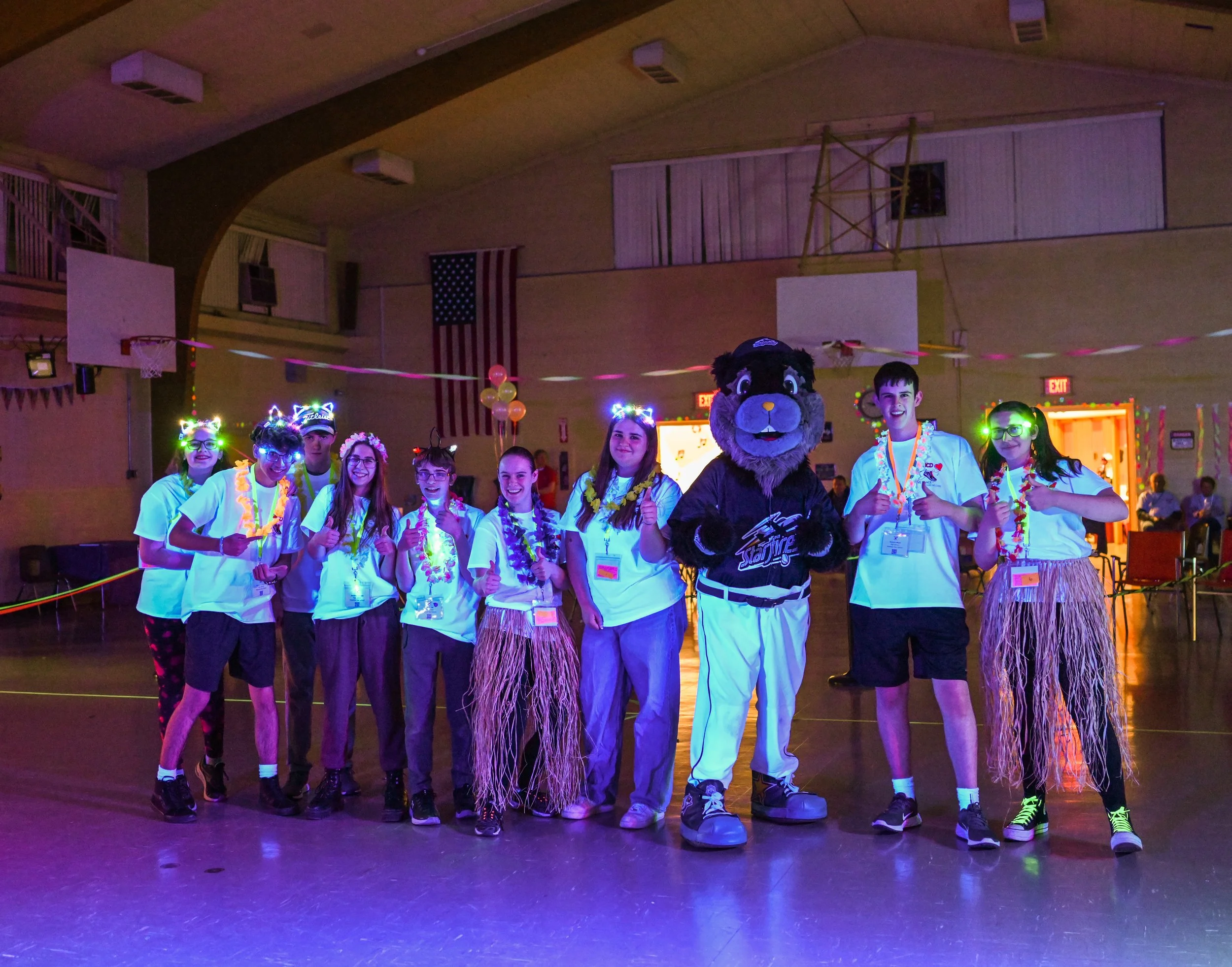 Group of young people with glow necklaces and accessories in a gymnasium, dressed with leis, coral skirts, and hats, with a mascot in the center, at a glow-themed party or event.
