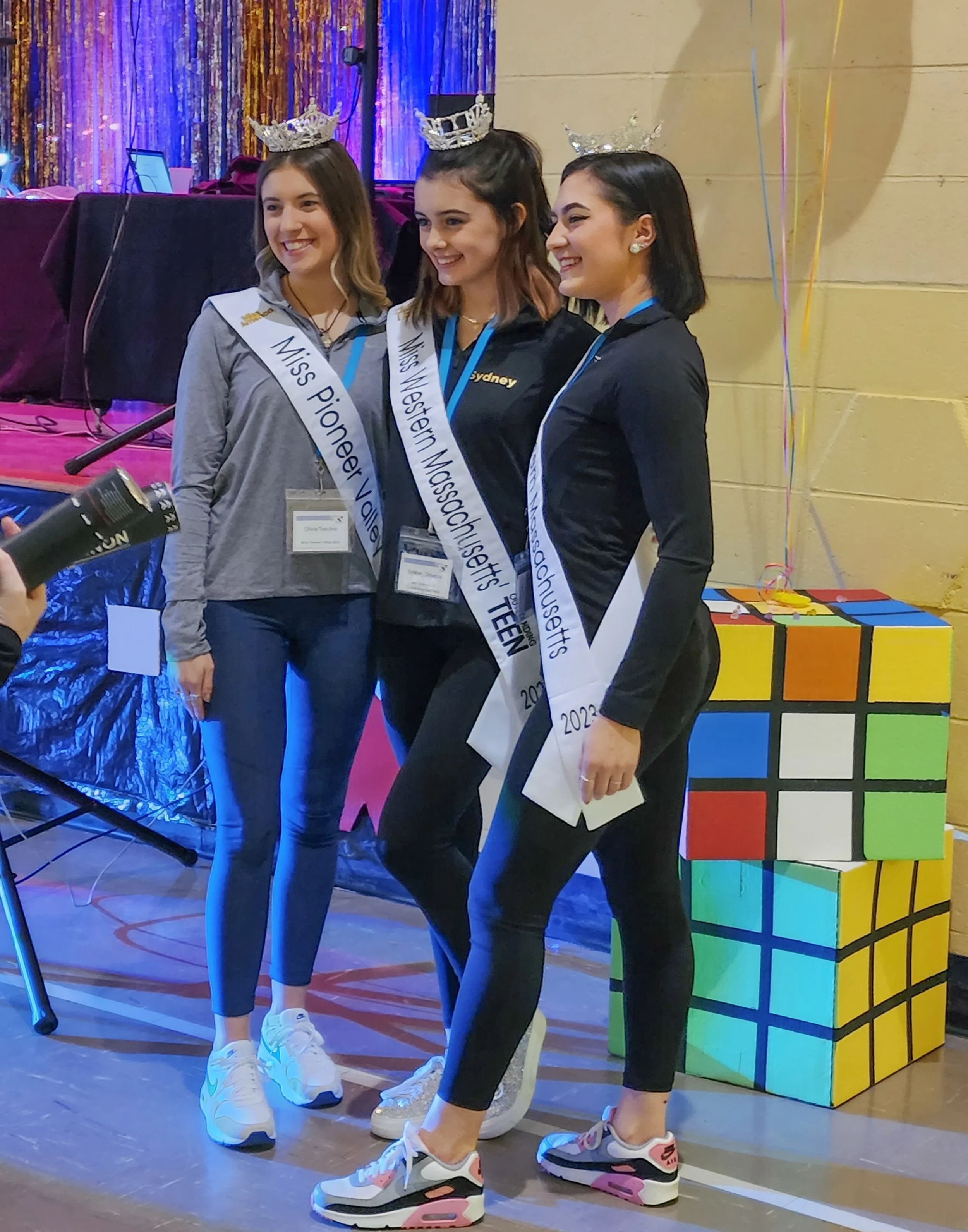 Three young women wearing crowns and sashes stand together at a pageant or competition. They are smiling and posing for a photo indoors, with colorful decorations in the background and a Rubik's cube stacked on the floor beside them.