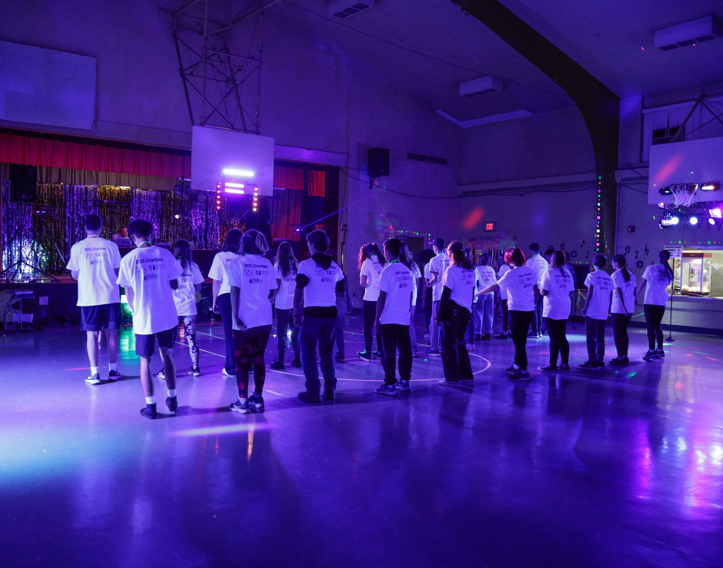 Students in white t-shirts standing in a line on a gymnasium floor under purple and blue stage lighting, with a decorated stage and disco-style lighting in the background.