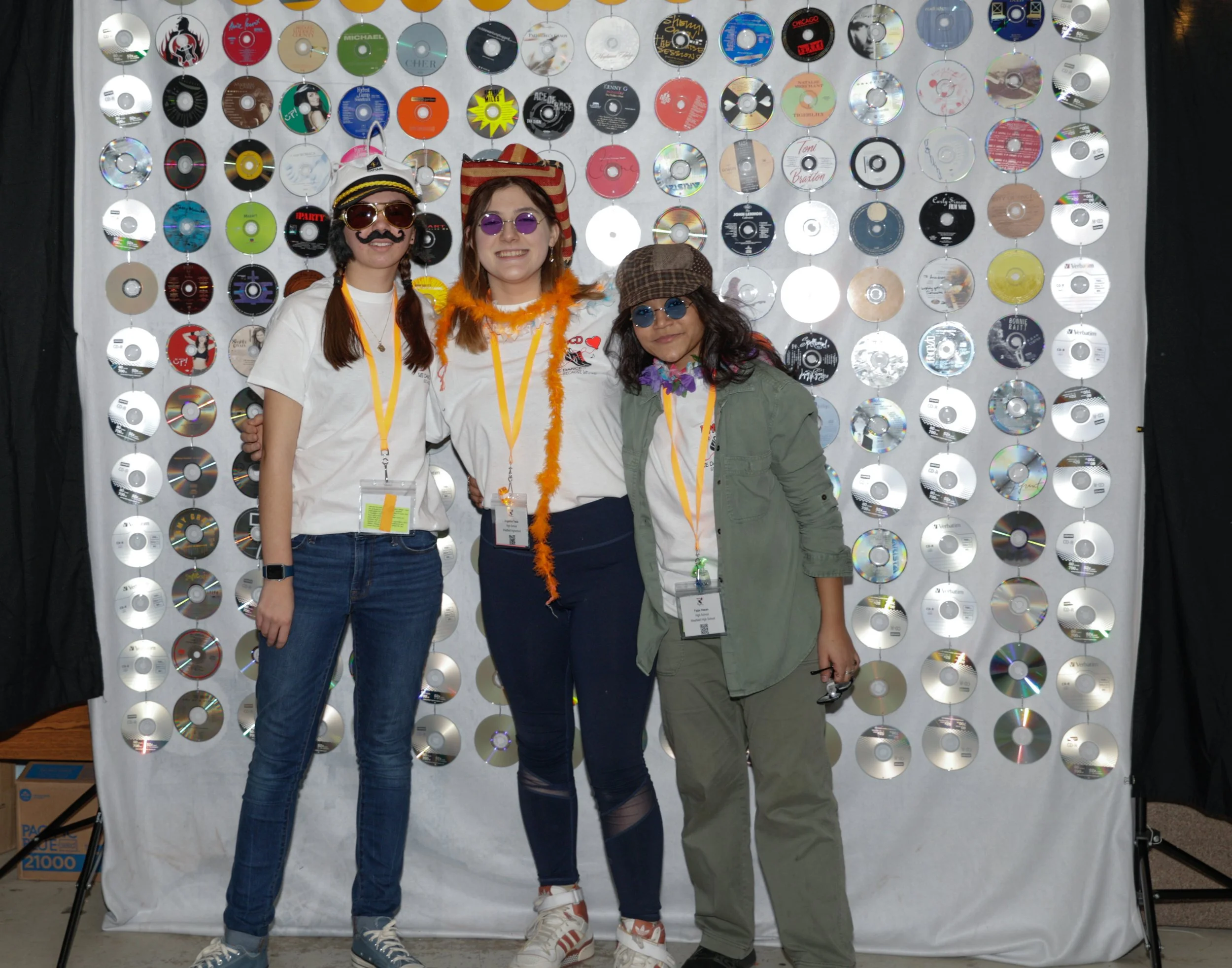 Three women dressed in casual clothing with fun accessories stand in front of a backdrop decorated with numerous compact discs. All are wearing lanyards with badges and sunglasses, smiling at the camera.