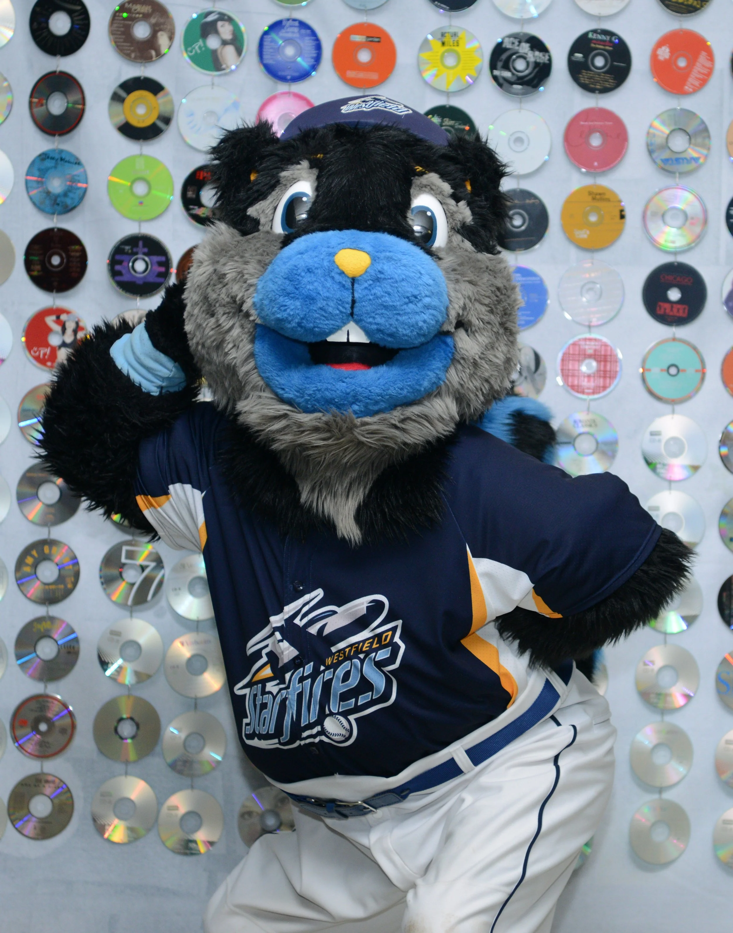 A person in a Westfield Westfield Jaffa Fire mascot costume posing with a hand on their head, standing in front of a wall decorated with colorful CDs.