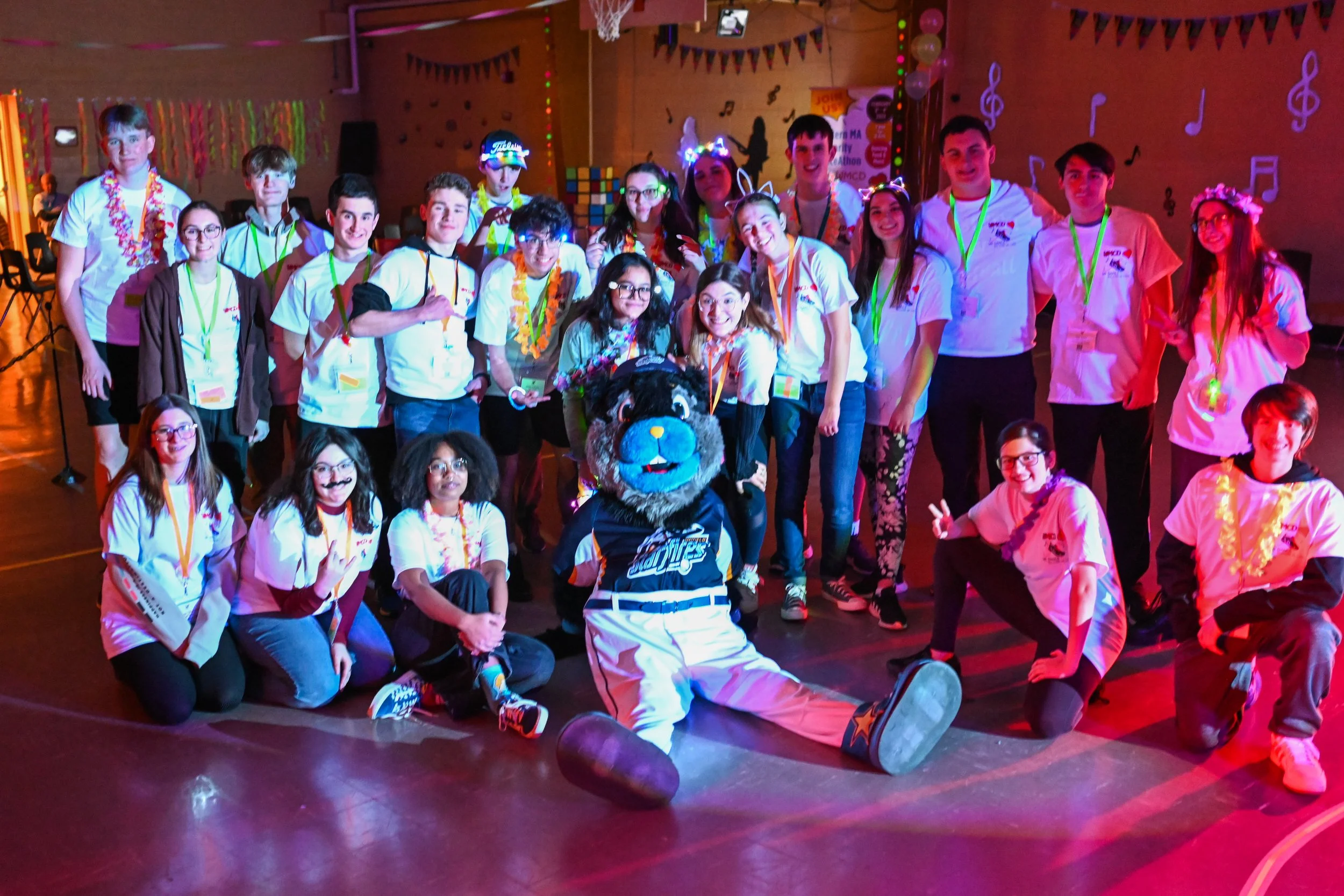 Group of young people and a mascot at a celebration or event, with colorful decorations including banners, balloons, and musical notes on the wall, in a gymnasium or hall.