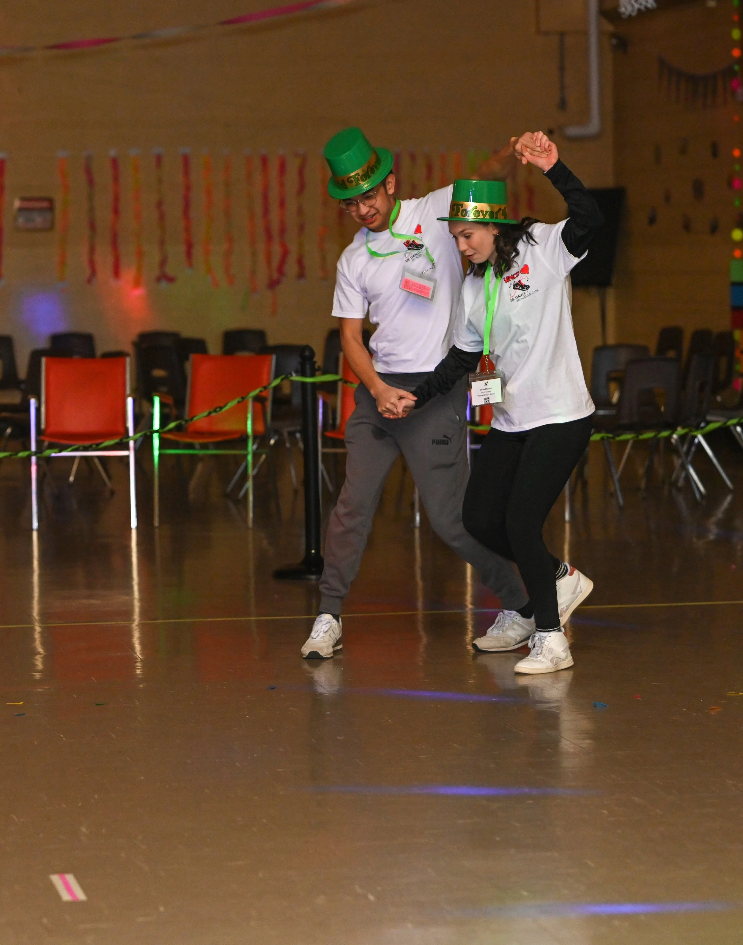 Two people dancing at a celebration, wearing green hats and party accessories, with chairs and colorful decorations in the background.