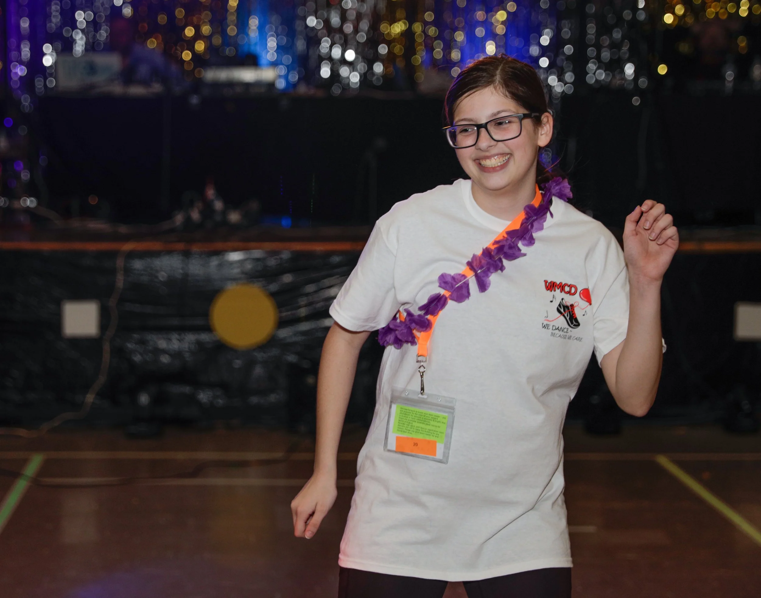 A young girl dancing in front of a dark backdrop with colorful blurred lights, wearing glasses, a white T-shirt with and a purple lei around her neck, and a green event badge hanging from her neck.