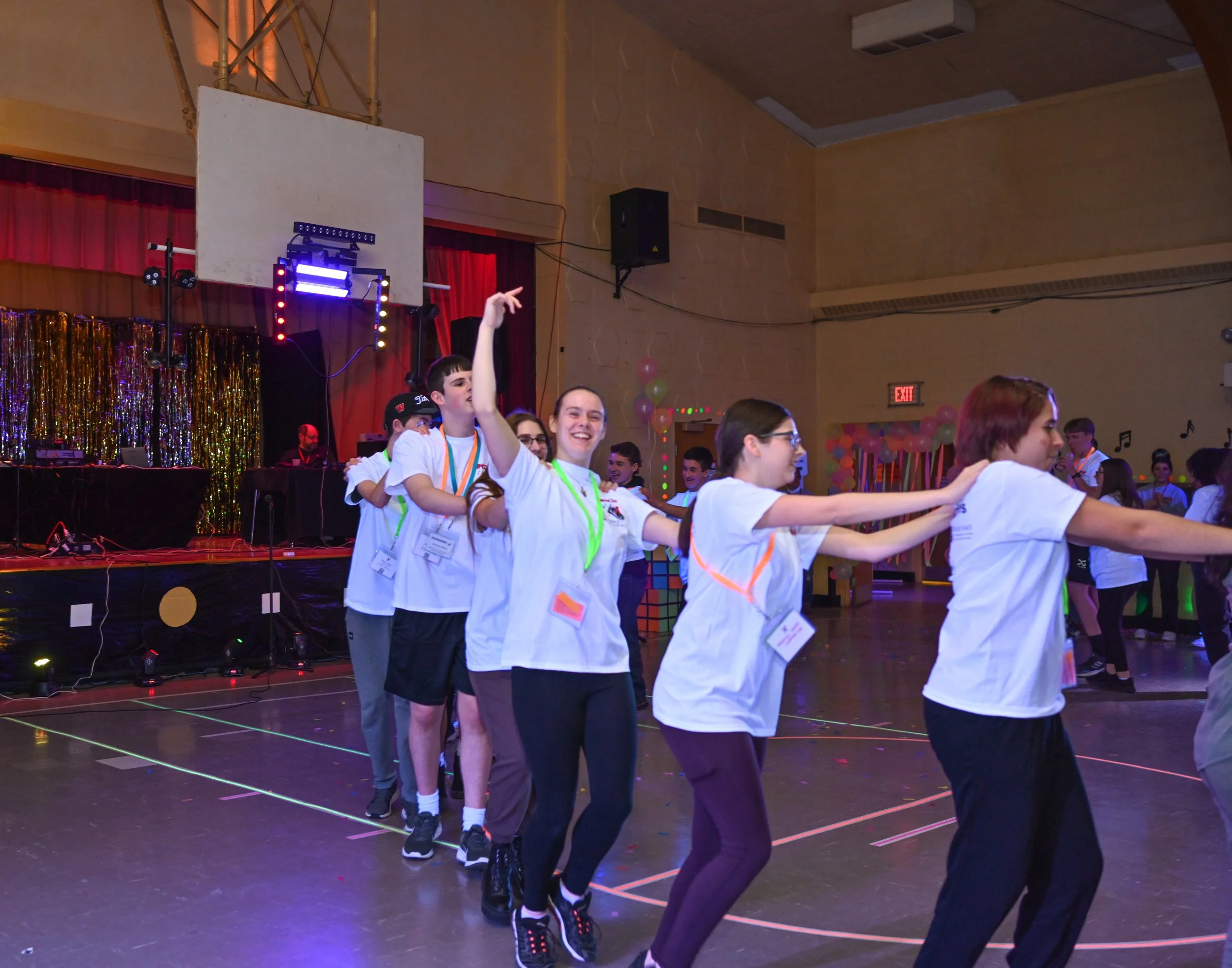 Group of young people at a dance party, standing in a conga line, inside a decorated hall with balloons, balloons, stage, and DJ setup in the background.