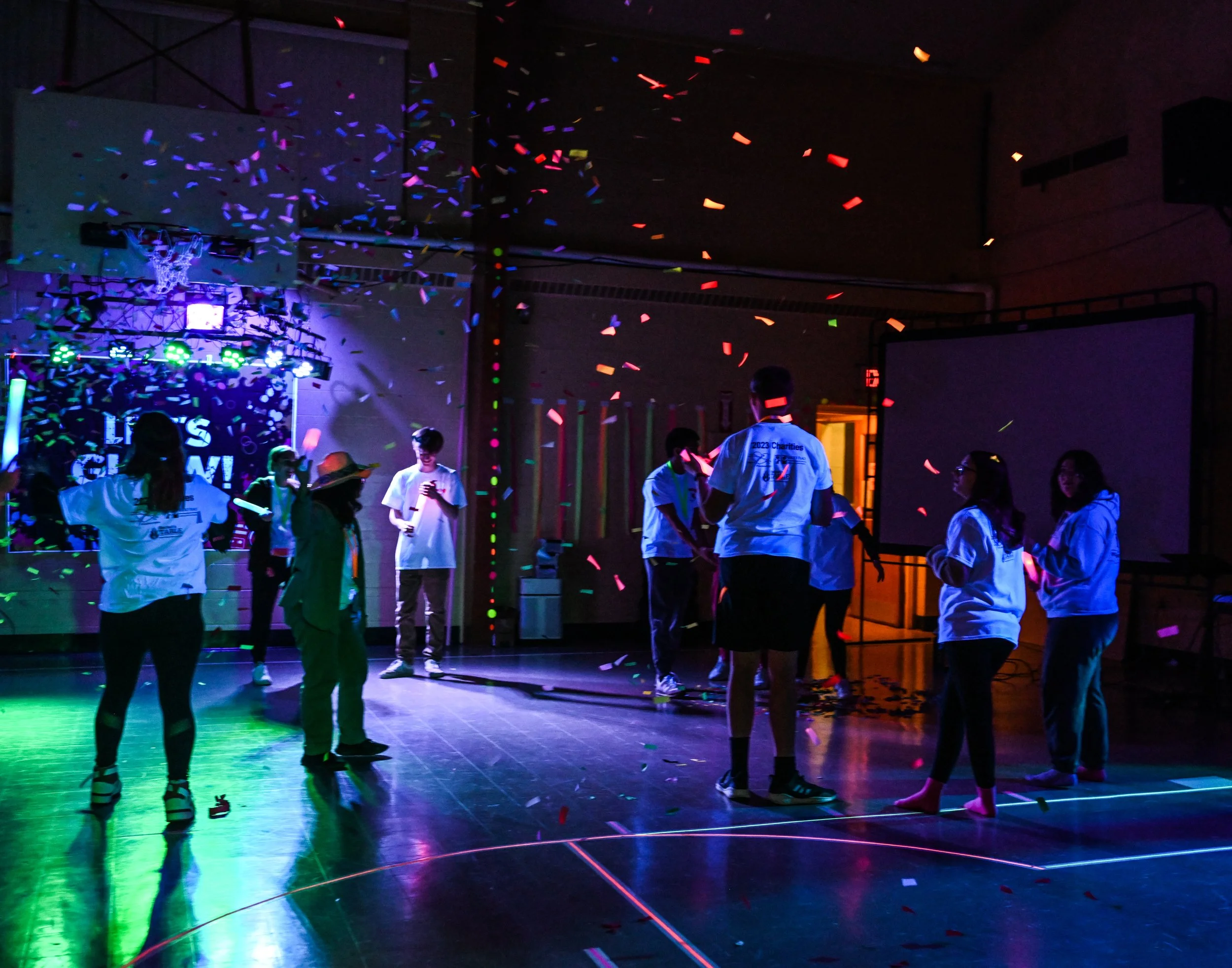 People celebrating with confetti in a gymnasium, illuminated with colorful party lights, some wearing white shirts and engaging in conversation.