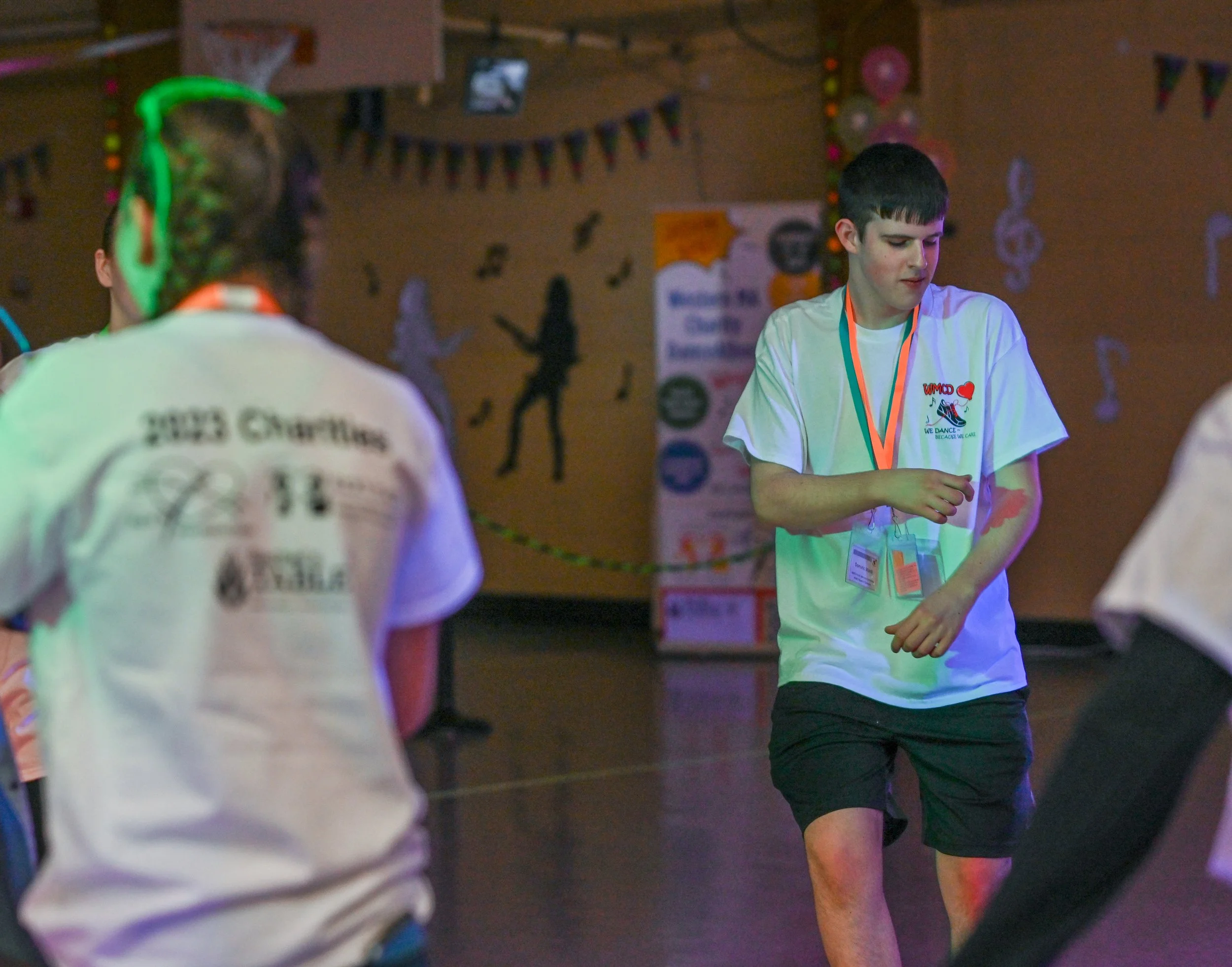 A group of young people at a dance event with a person in front wearing a white t-shirt with logos, another boy in the center with a white t-shirt and shorts, and a decorated background with music notes and silhouettes.