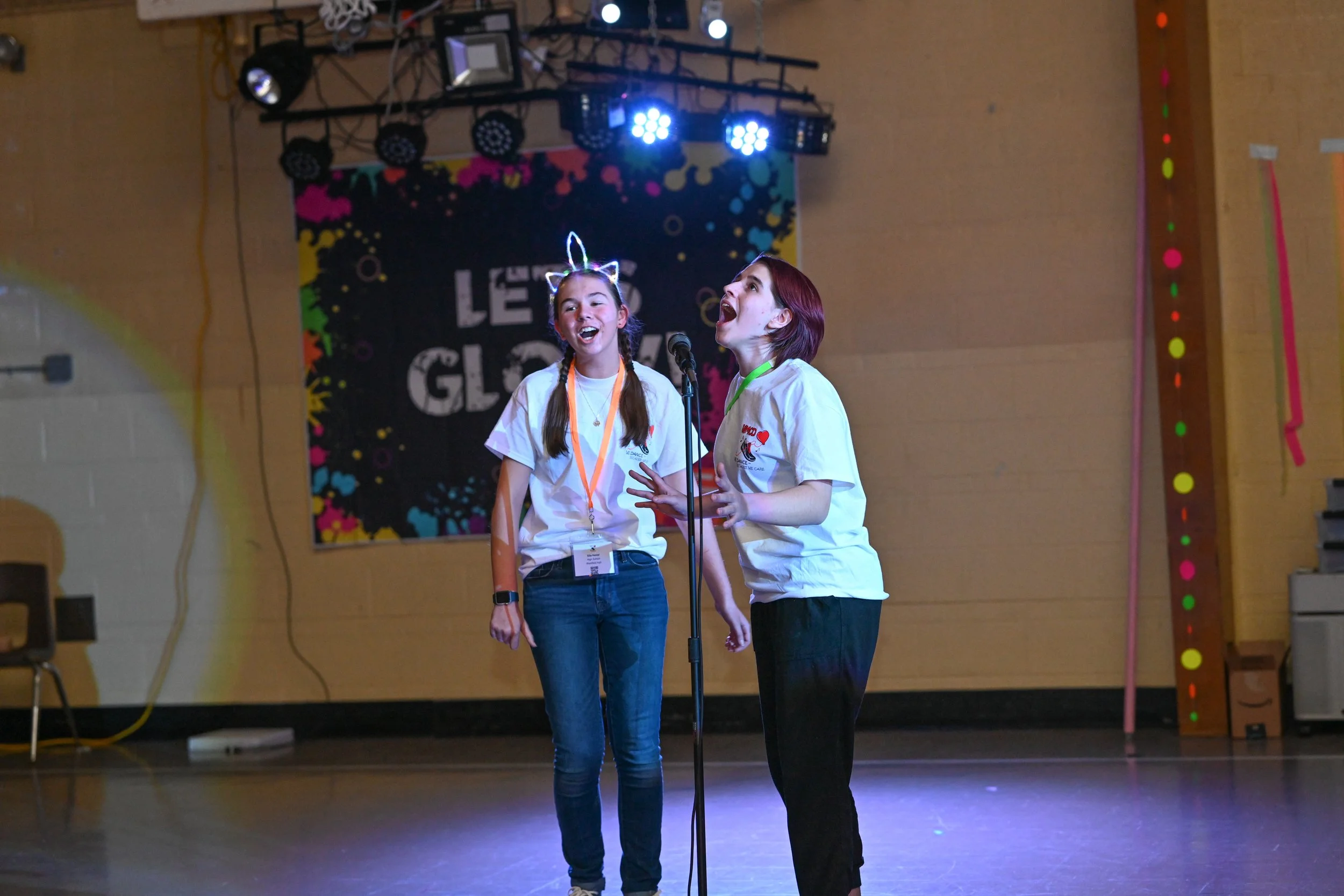 Two young girls singing on stage during a performance at an event, with one girl wearing a glowing cat ear headband. They are standing in front of a microphone with colorful stage lights above and a backdrop that reads 'LE GAL' with a colorful design