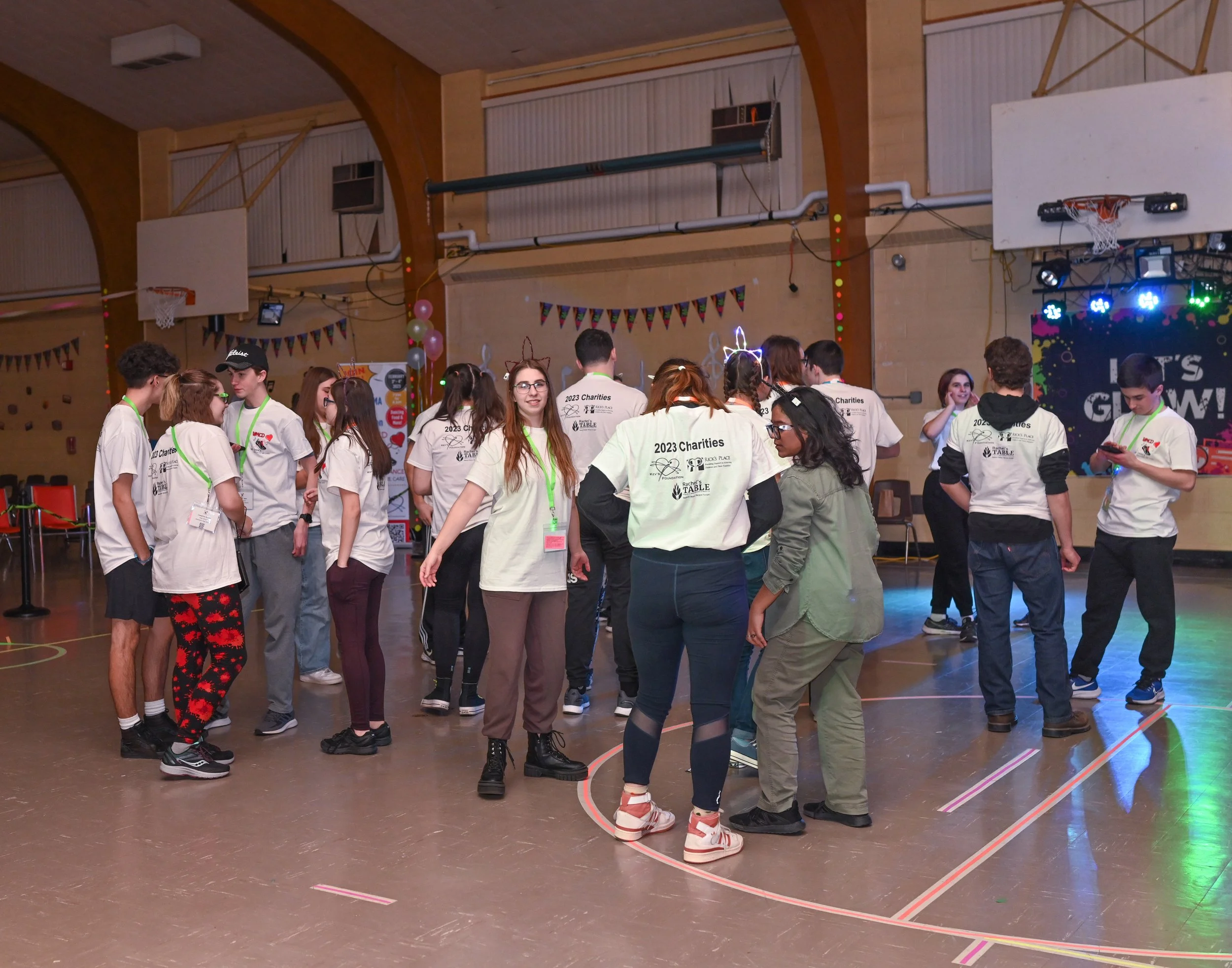 Group of teenagers in a gymnasium during a charity event, wearing matching white t-shirts with '2023 Charities' printed on the back, some with festive light headbands, standing and chatting under basketball hoops with a colorful stage backdrop readin