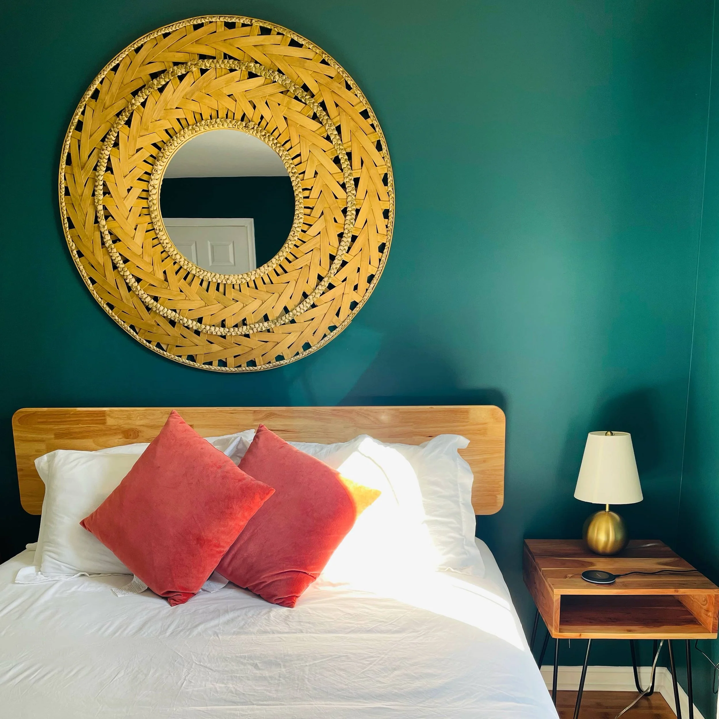 A bedroom with a green wall, a wooden headboard, two red cushions, white bedding, a wooden nightstand with a gold lamp, a smartphone, and a large round decorative mirror with wooden patterns hanging above the bed.