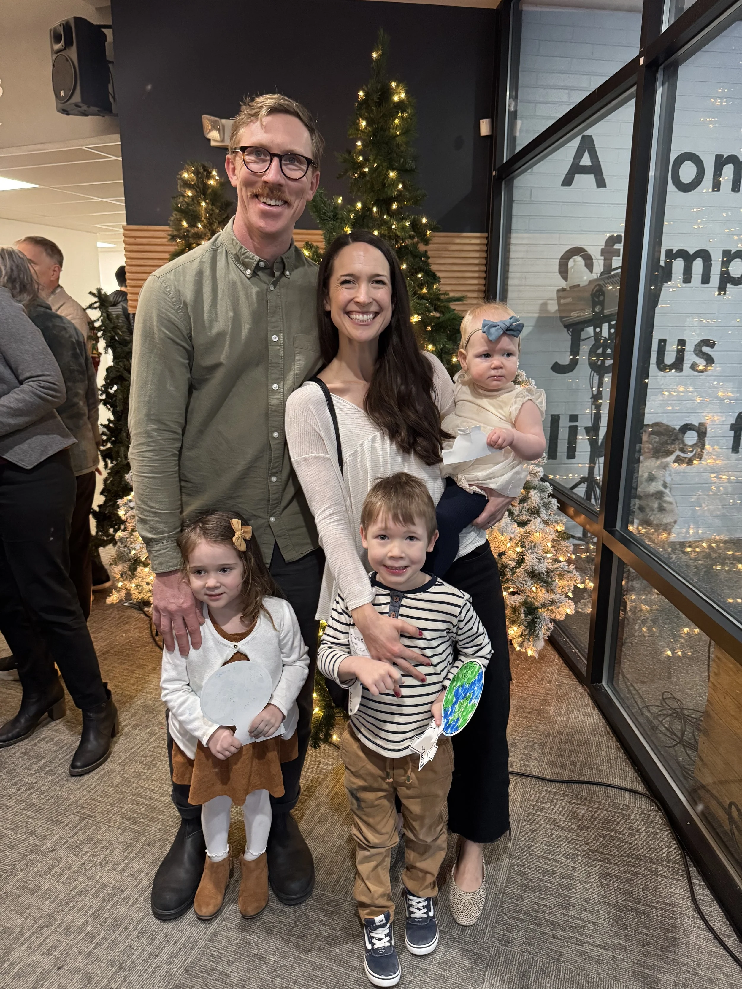 A group of four children and two adults standing in front of a decorated Christmas tree indoors. Behind them are other people, and the window on the right shows part of a sign with visible letters.