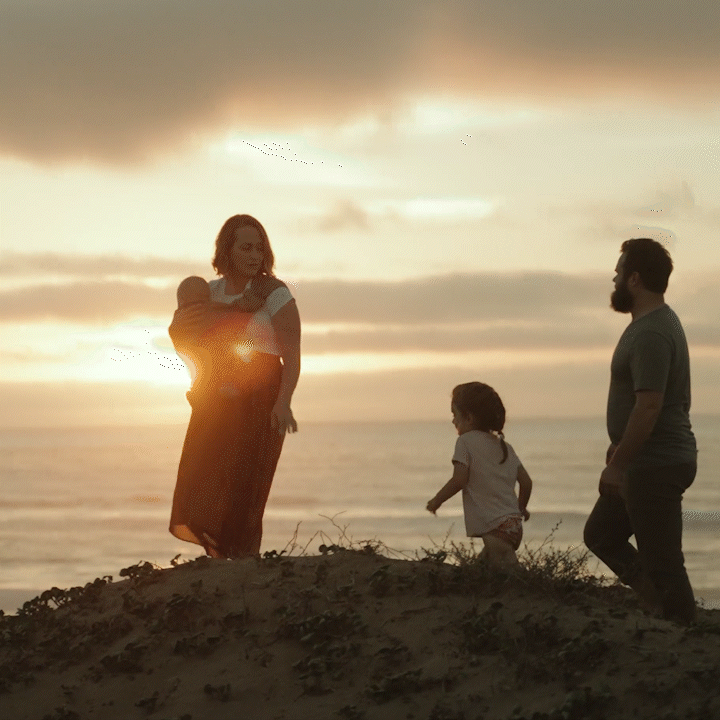 A family of three on a beach at sunset: a woman holding a baby, a young girl, and a man standing nearby, with the sun setting over the ocean in the background.