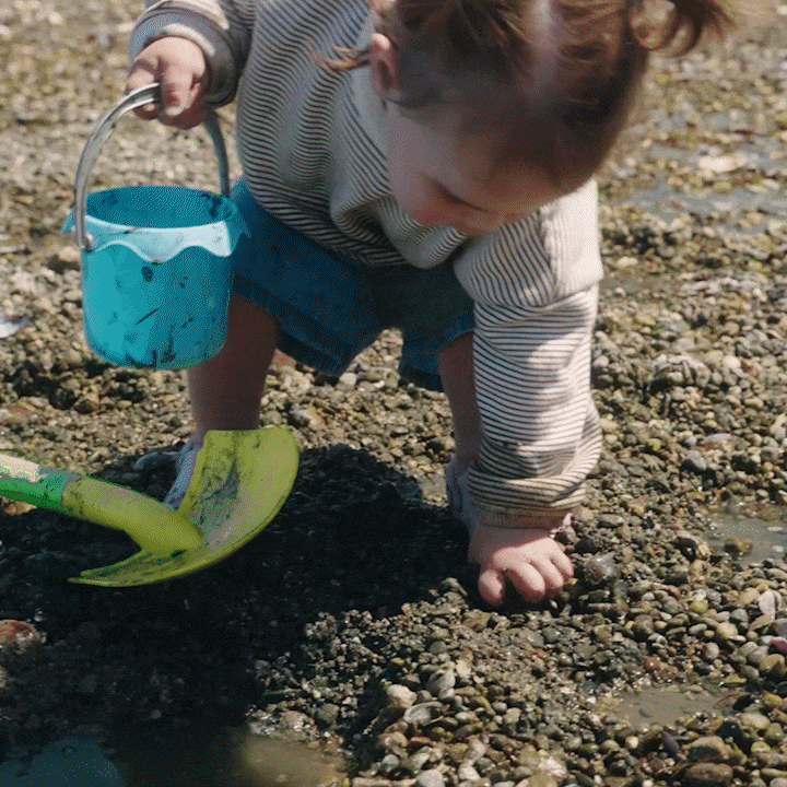 A young child crouches on a pebbled ground, holding a small blue bucket with a metal handle in one hand and a green shovel in the other, exploring outdoors.