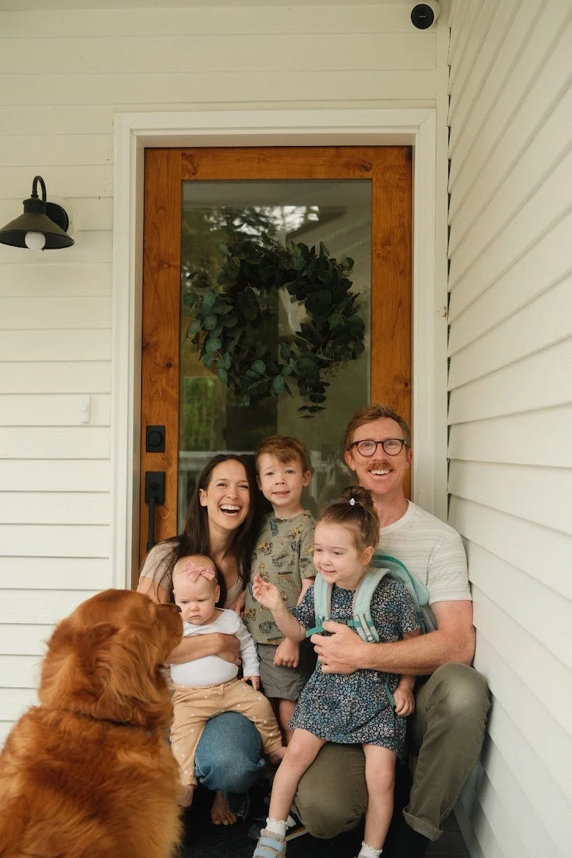 A smiling family of four with a dog on a porch in front of a white house with a wooden door and a large green wreath.