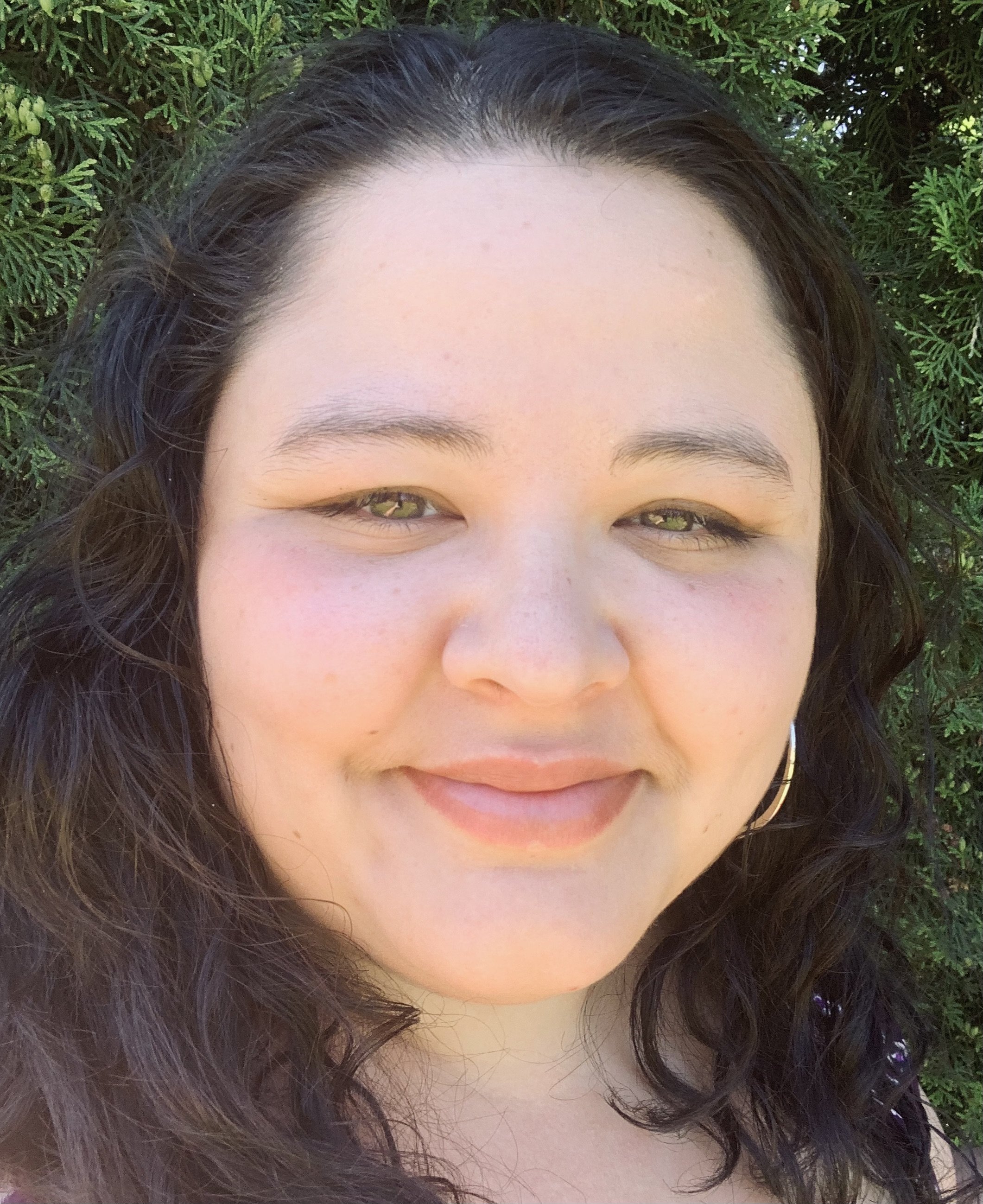 Mercedes is smiling warmly in front of a hedge on a sunny day.