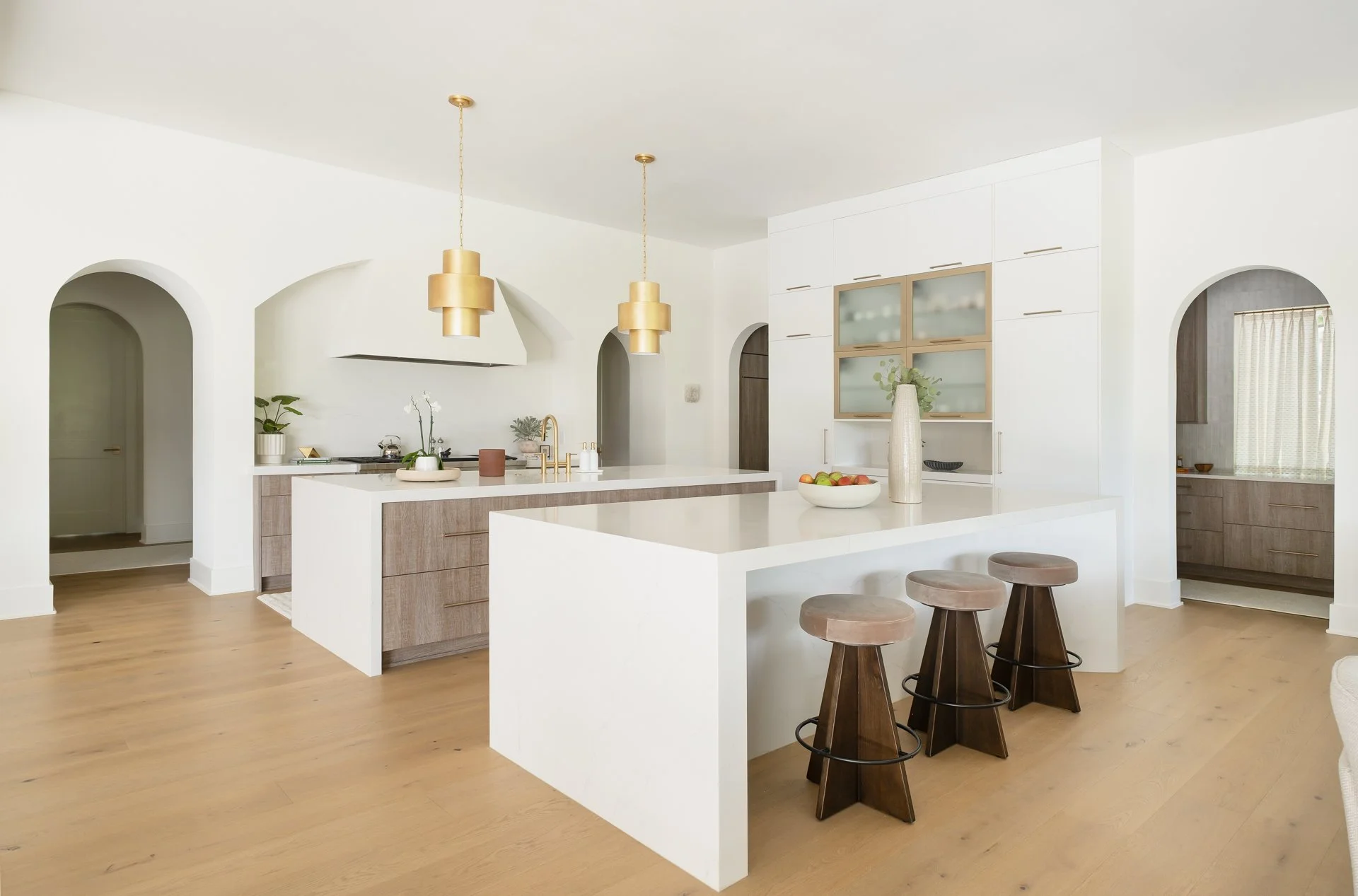 Modern kitchen with white cabinets, gold pendant lights, wooden stools, and a central island with decorative vases and a bowl of fruit.