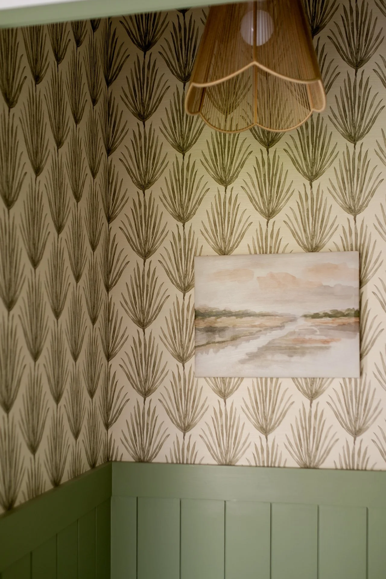 Corner of a room with patterned wallpaper featuring large green leaves, a landscape painting of a river scene, and a pendant lampshade hanging from the ceiling.