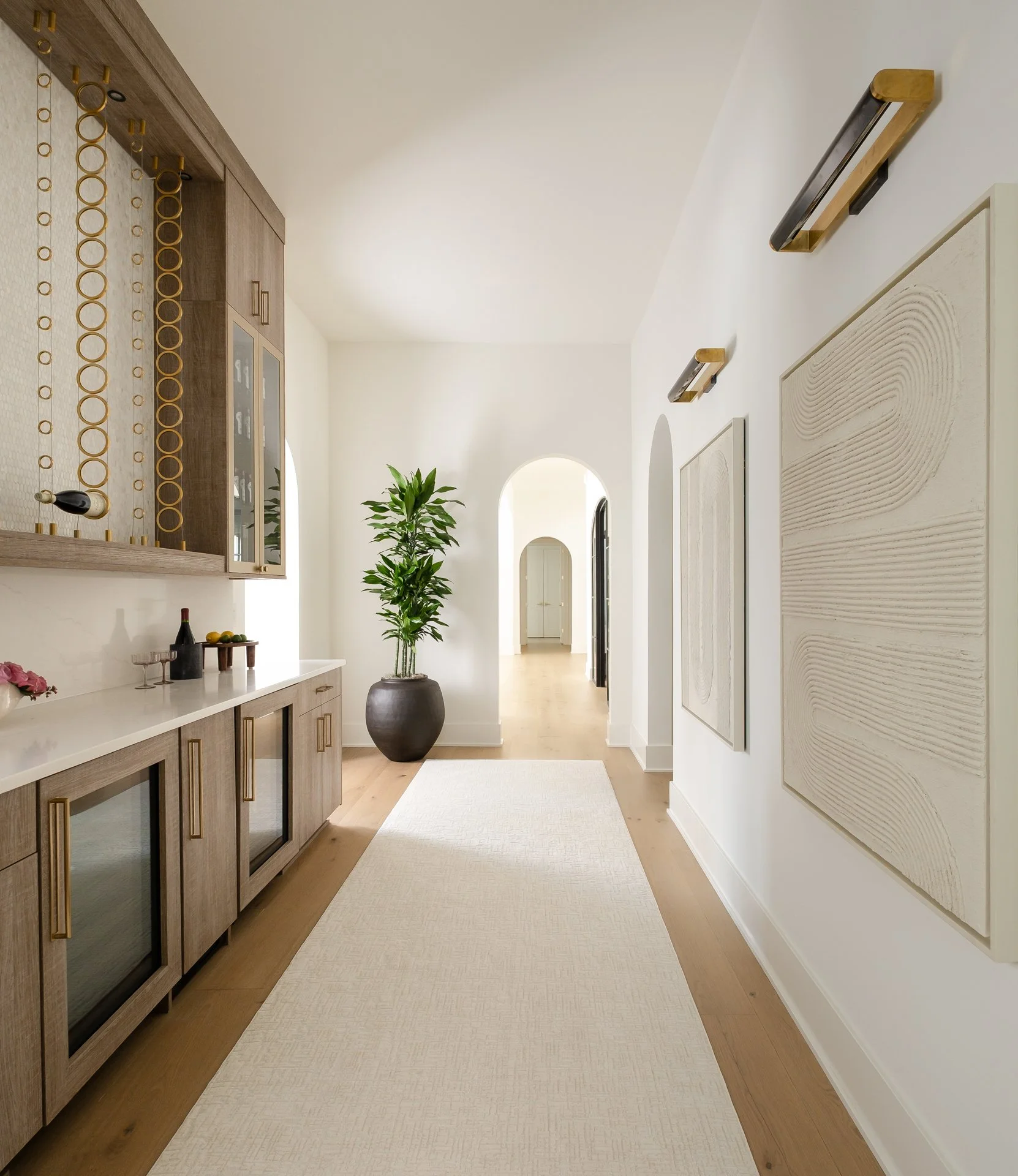 Bright hallway with light wood flooring, white walls, modern wall art, and potted plant, leading to a doorway in a contemporary home.