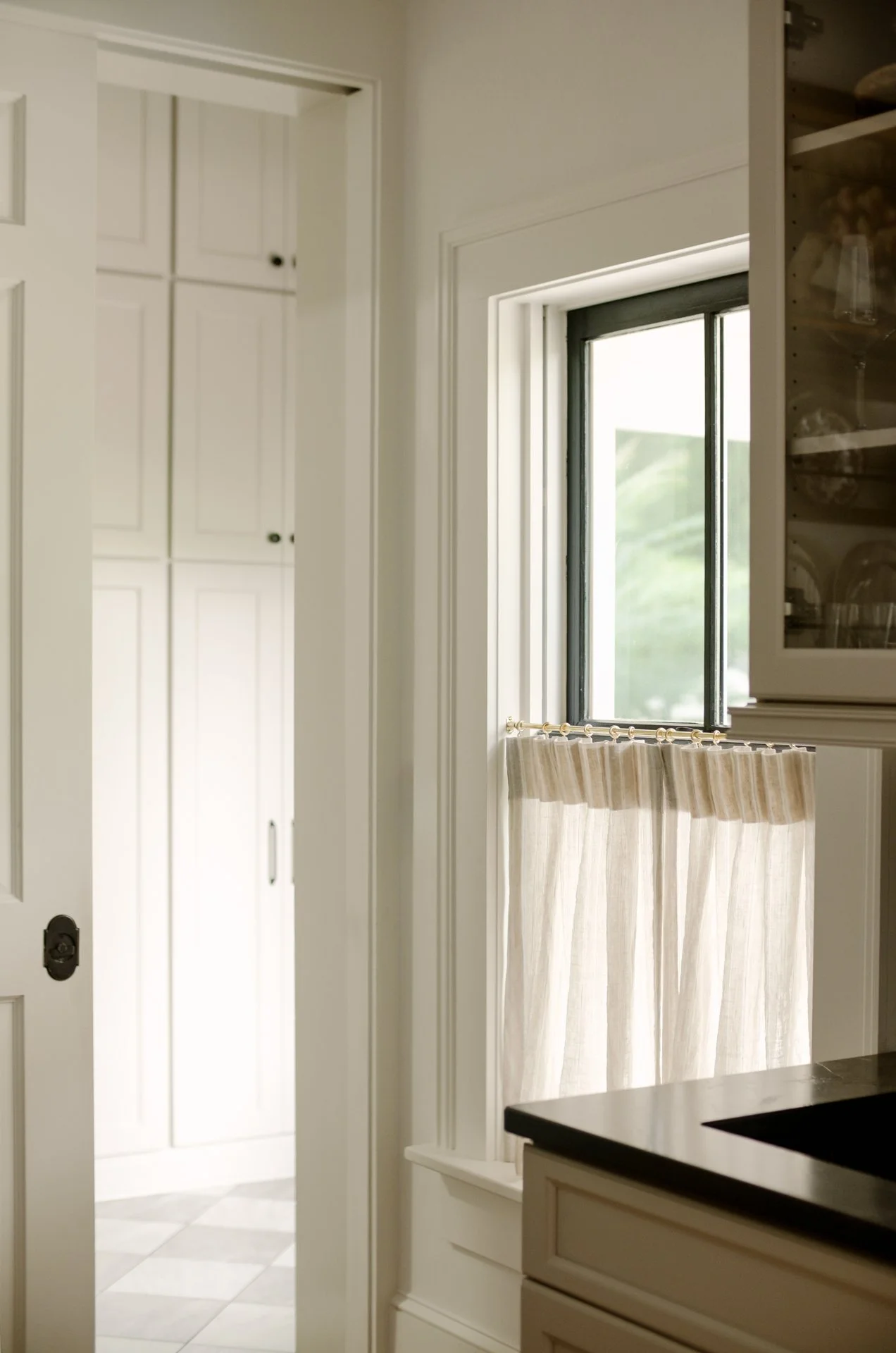 Kitchen window with a beige curtain, adjacent to a cabinet with glass doors, and a white pantry closet with black knobs in the background.