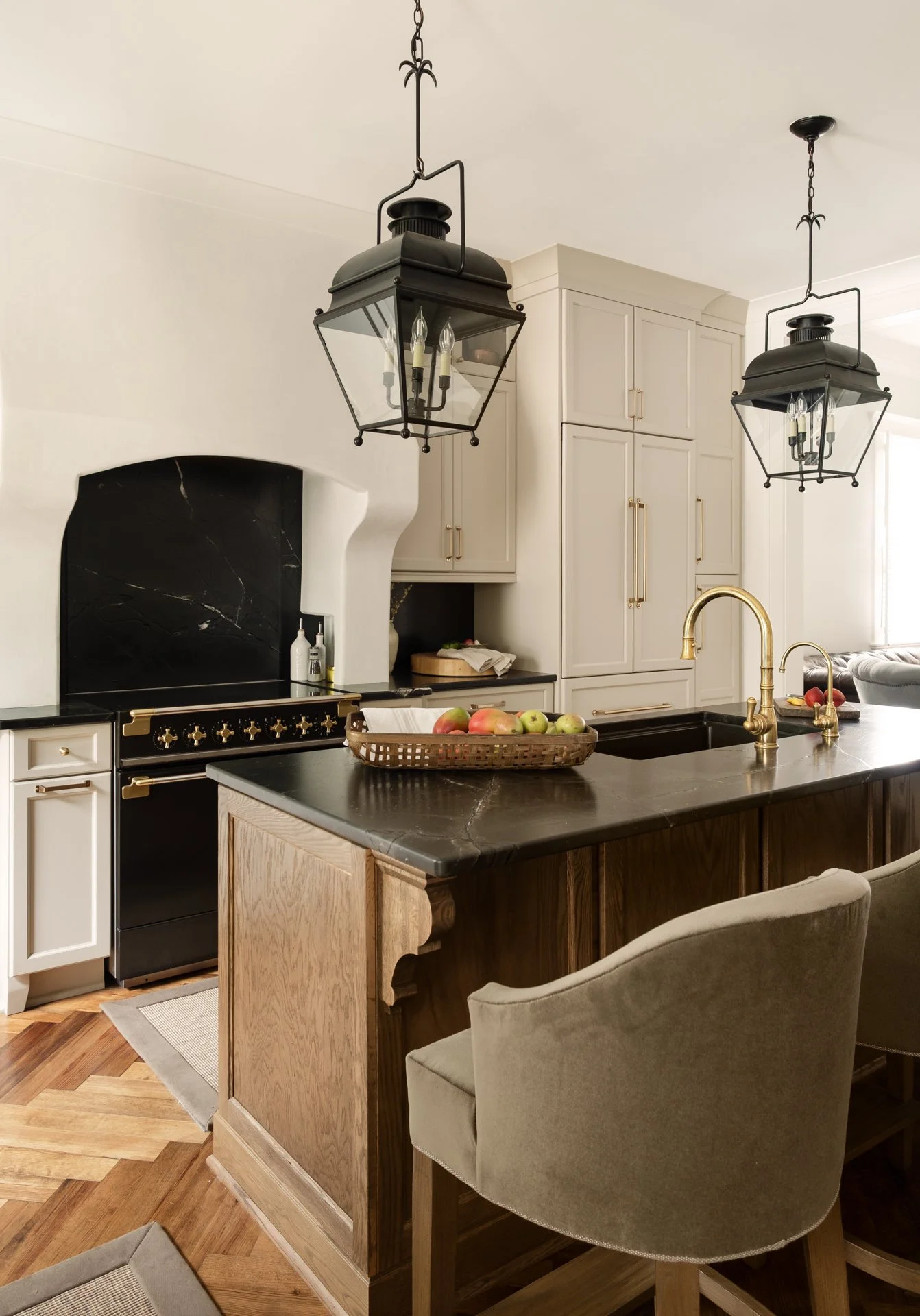 Modern kitchen with a black marble countertop, white cabinetry, and brass fixtures. Two hanging lantern-style lights are above the island, which has a basket of apples and a double brass sink. A black stove with brass accents is in the background, an