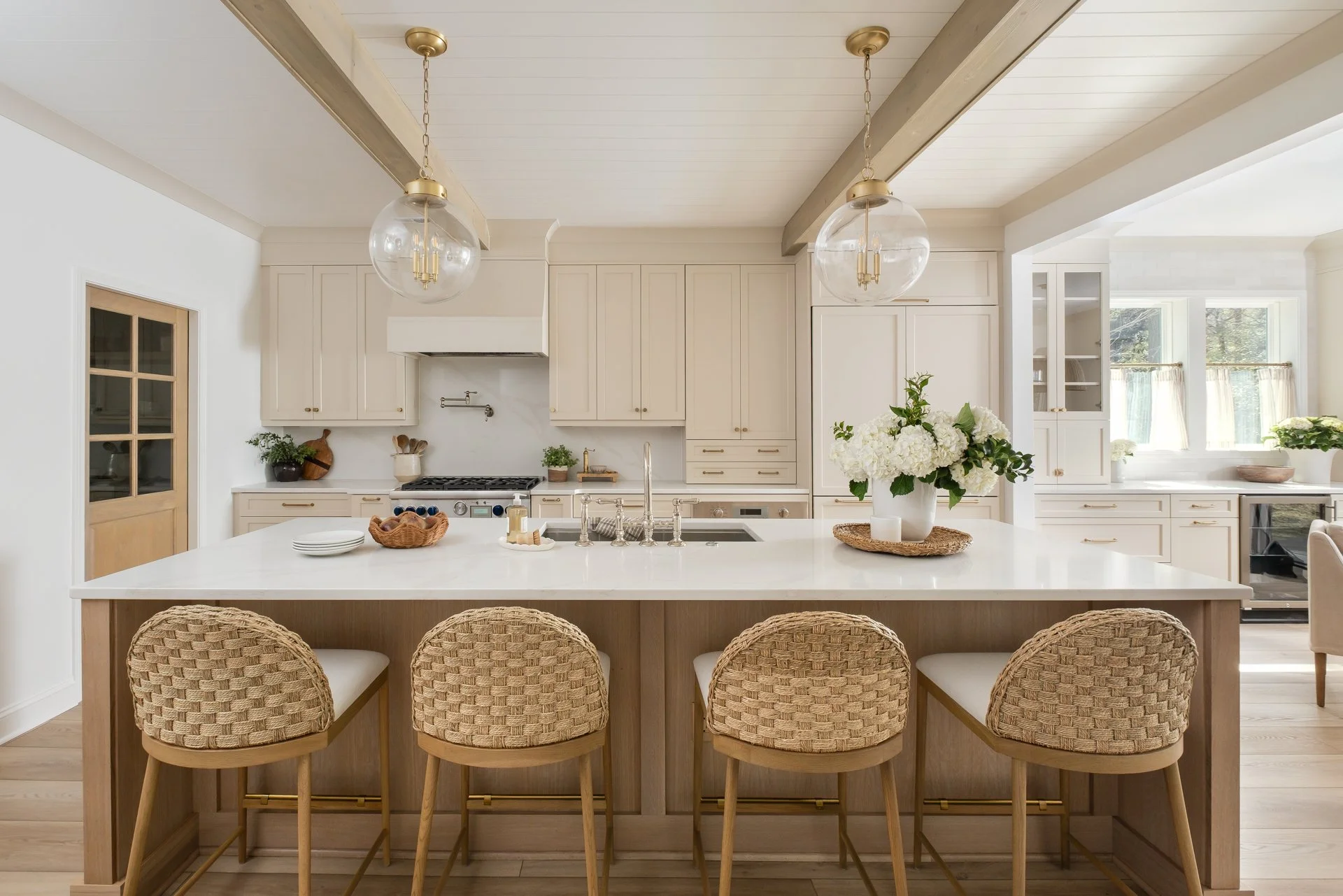 A bright and airy kitchen with an all-white color scheme, featuring a large island with a white countertop and four beige woven bar stools. The kitchen has white cabinets, a stove, and a window with natural light. There are hanging pendant lights wit