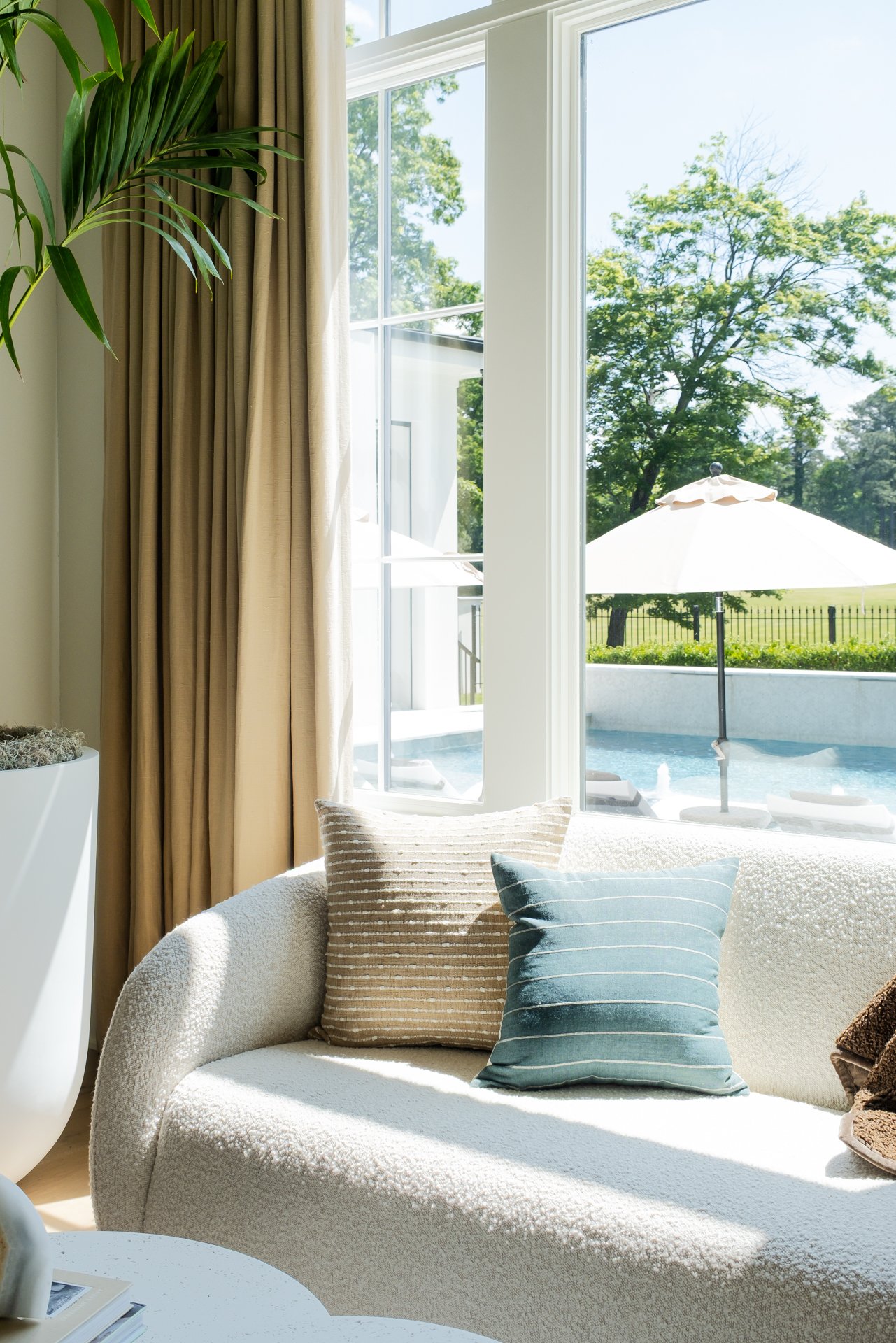Living room with a large window overlooking a pool and garden, beige curtains, cream textured sofa with throw pillows, and a side table.