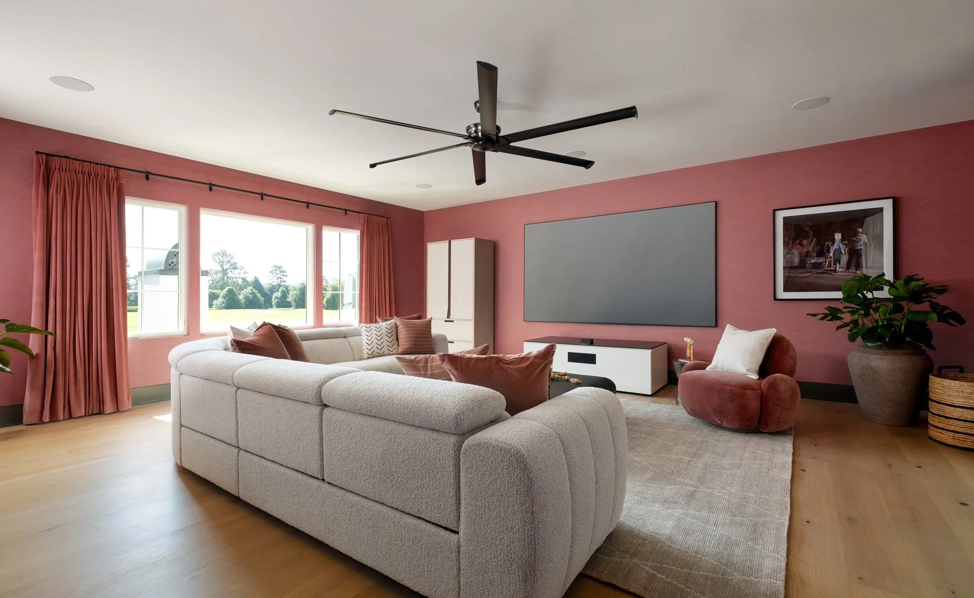 Living room with pink walls, large white sectional sofa with brown and white pillows, a pink velvet chair, potted plant, large windows with pink curtains, wall-mounted TV, and a ceiling fan.