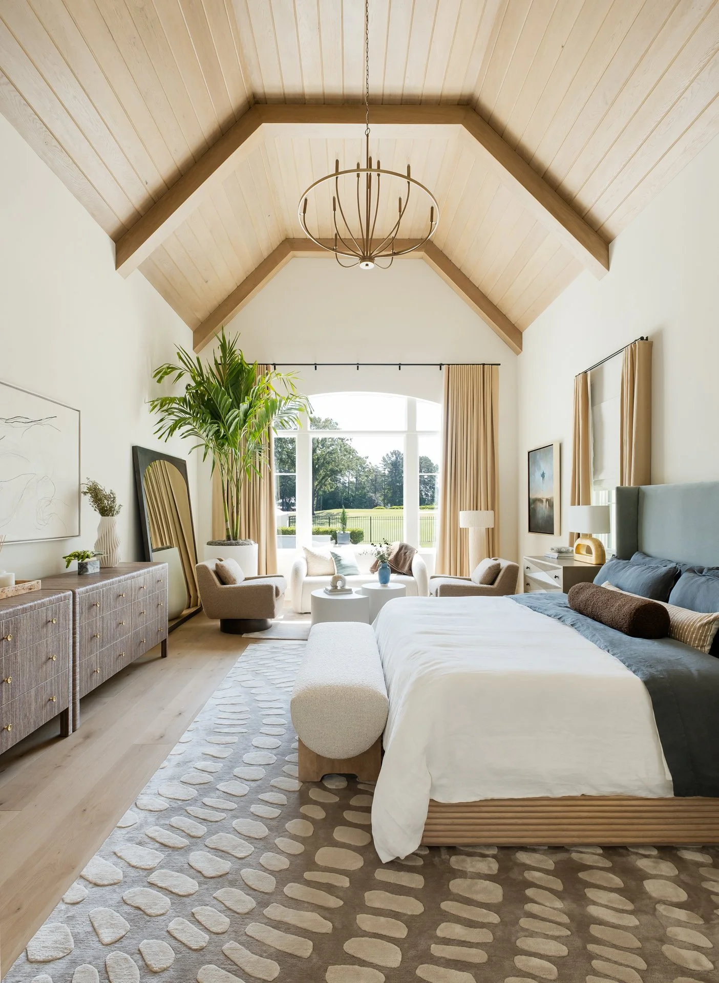 Bright bedroom with vaulted wooden ceiling, large window with curtains, and modern decor. Bed with pillows, a patterned rug, a seating area with chairs and a table near the window, and a large potted plant.