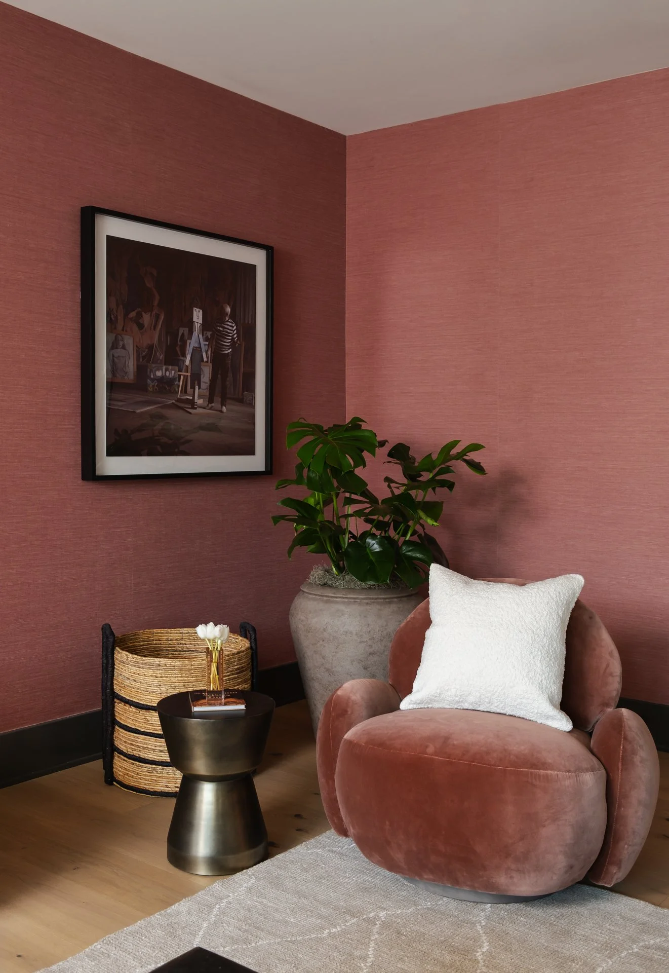 A cozy corner with a pink velvet armchair, a white pillow, a small round black side table, a large potted plant, a framed artwork on a pink textured wall, a basket, and a small vase with white flowers.