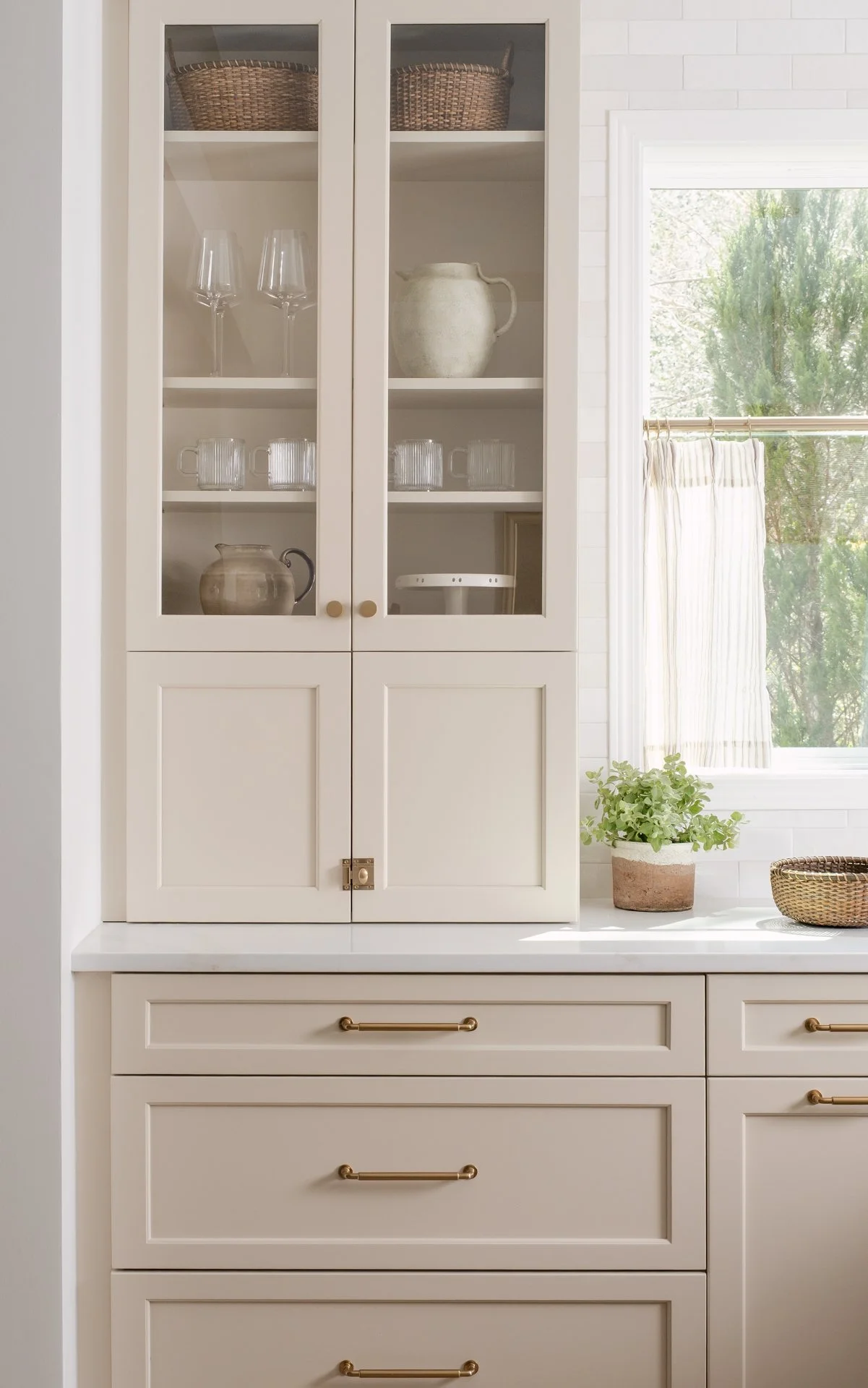 A cream-colored kitchen cabinet with glass doors, displaying glassware and pottery inside, next to a window with a plant on the countertop.