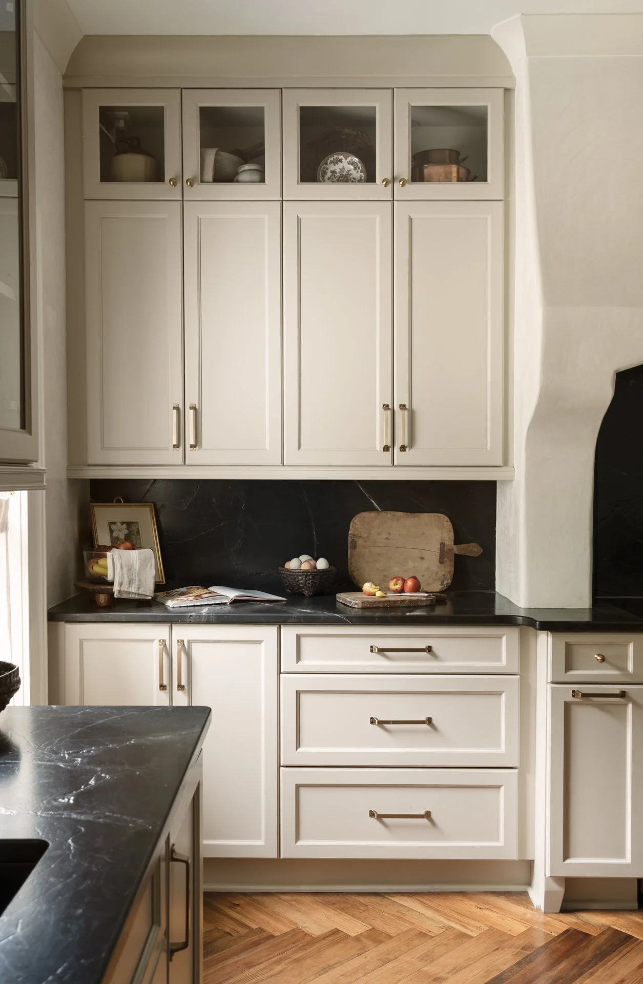 A kitchen with white cabinets, black countertops, and a wooden cutting board with apples and eggs. There are bowls and dishes in the upper cabinets, a framed picture, a bowl of bananas, and a towel on the counter.