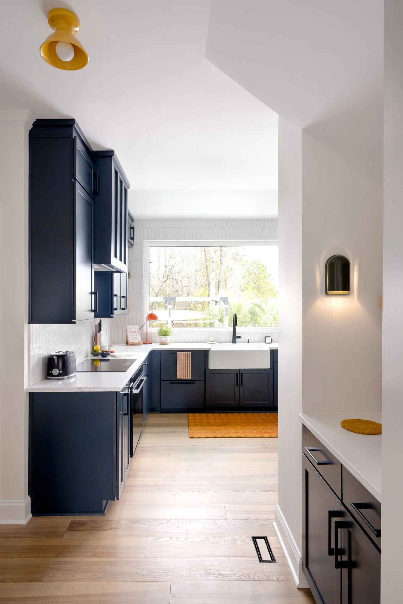 A modern kitchen with dark navy cabinets, white countertops, a large window, and light wooden floors.