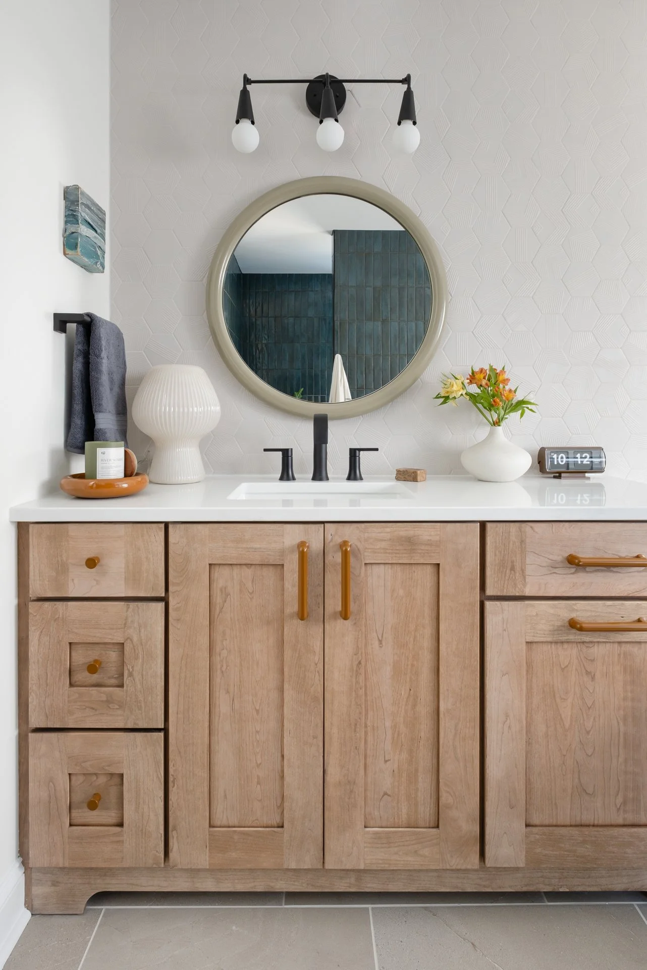 A modern bathroom vanity with a light wood finish, a white countertop, black faucet, round mirror, and decorative items including a vase with flowers and a clock.