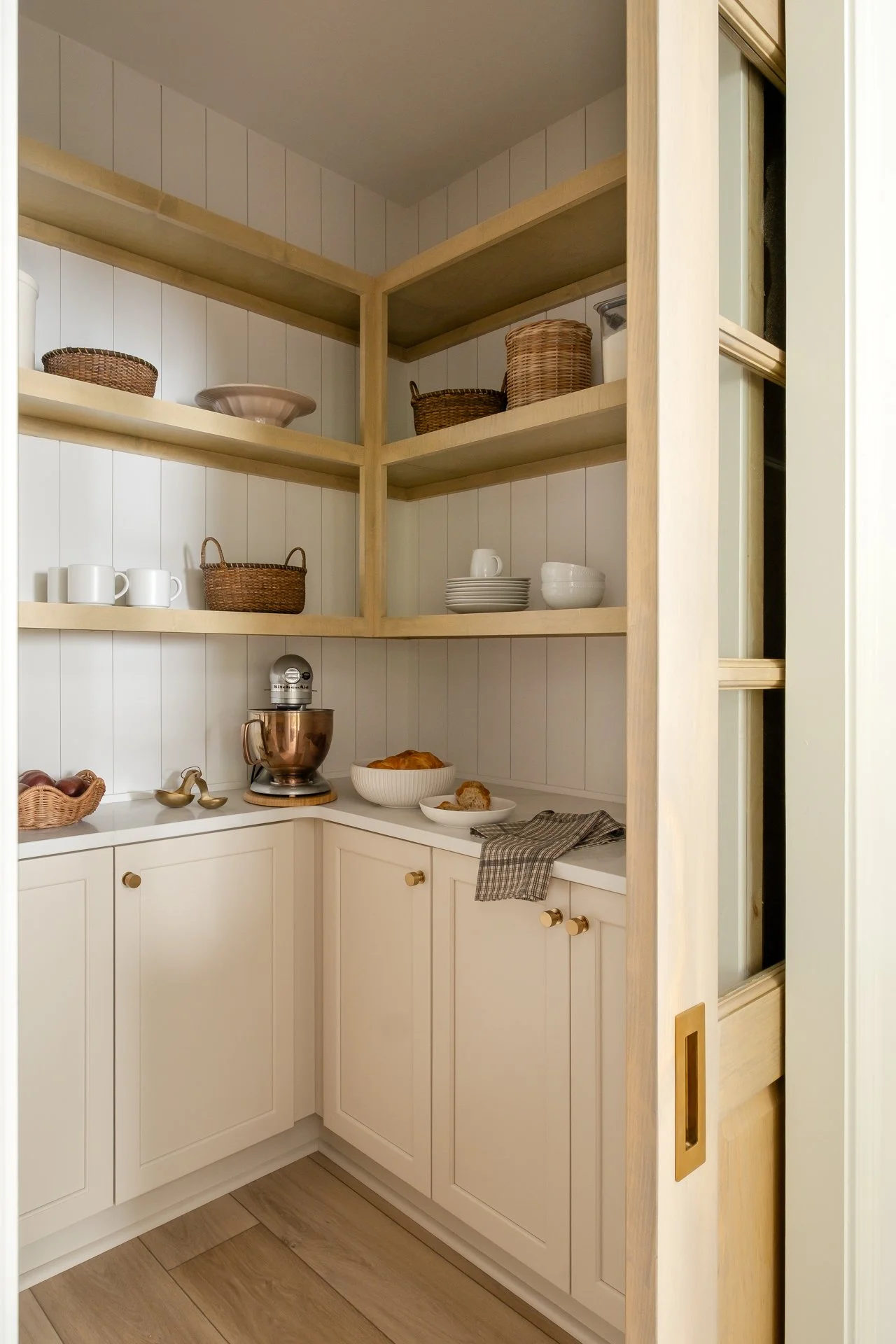 Kitchen corner with open wooden shelves filled with dishes, baskets, and bowls, white cabinets, and a countertop with a stand mixer, bread, and a cloth.
