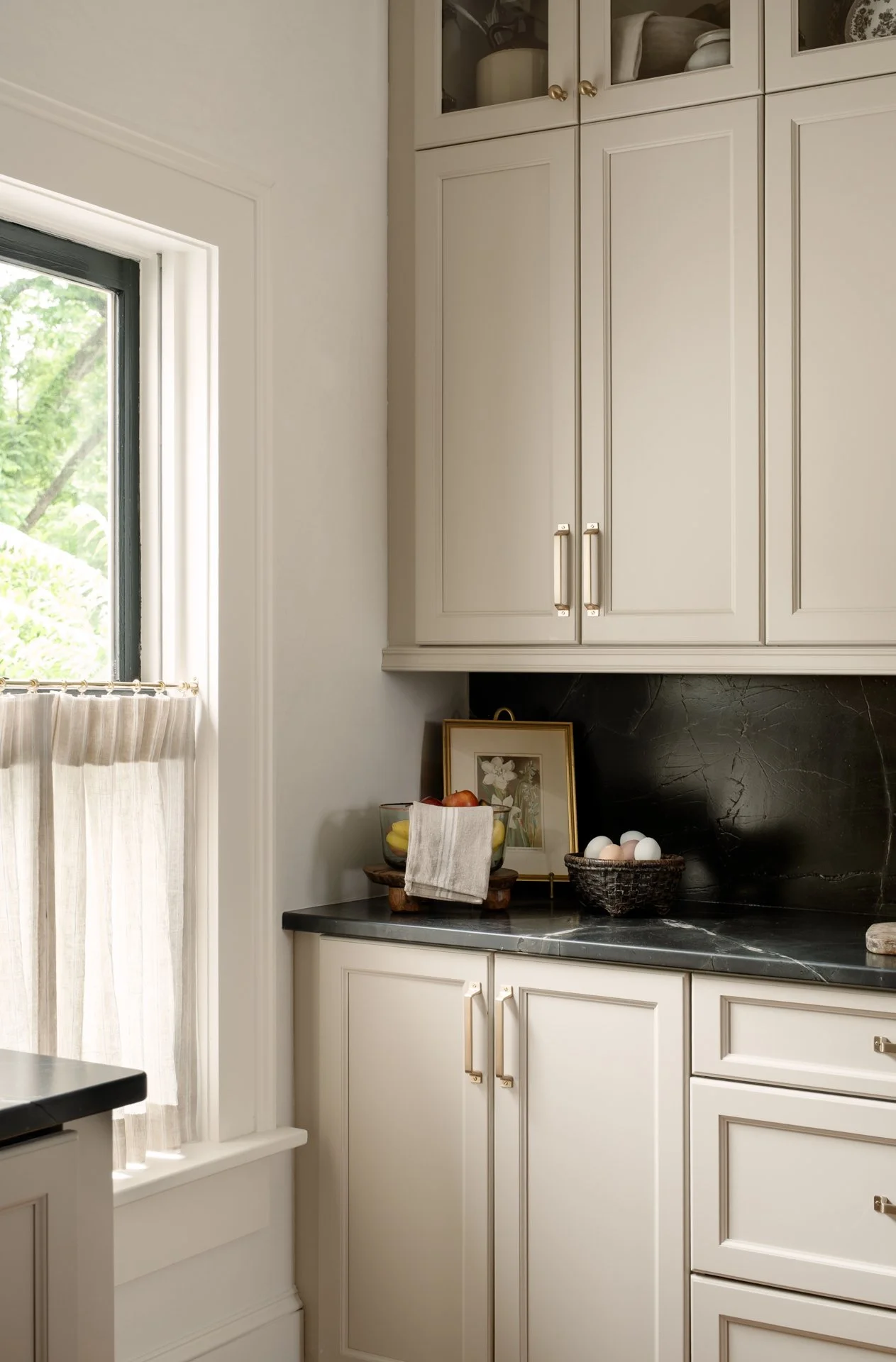 A kitchen corner with cream cabinets, black countertop, window with a beige curtain, and a small decor with eggs, fruit, and framed art.