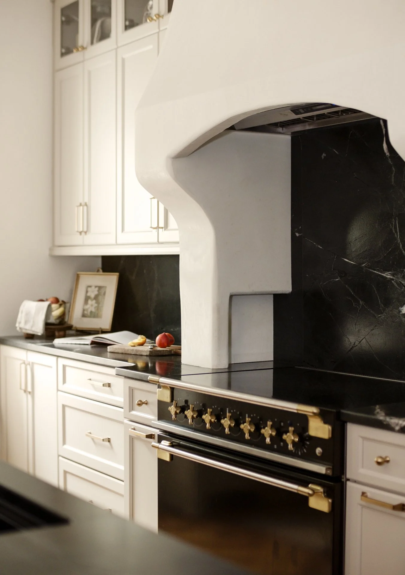 Kitchen with white cabinets, black stone countertops, and a black and gold stove. There are apples on the countertop and a framed picture leaning against the wall.